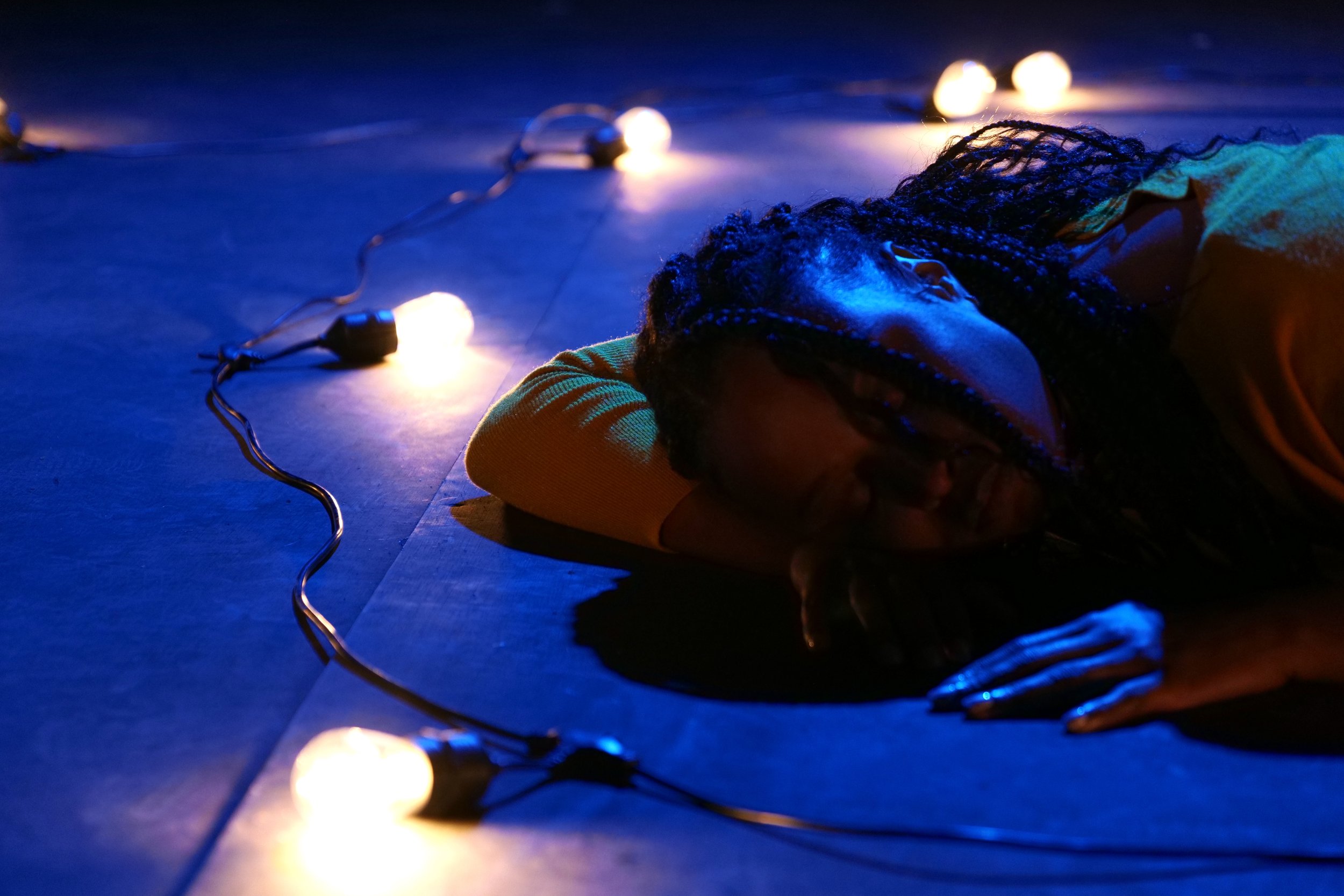  Photo Credit: Ryd Cook  A black woman lies on the floor, head turned to the side toward the viewer. In front of her, a string of lightbulbs illuminate the floor around her. 