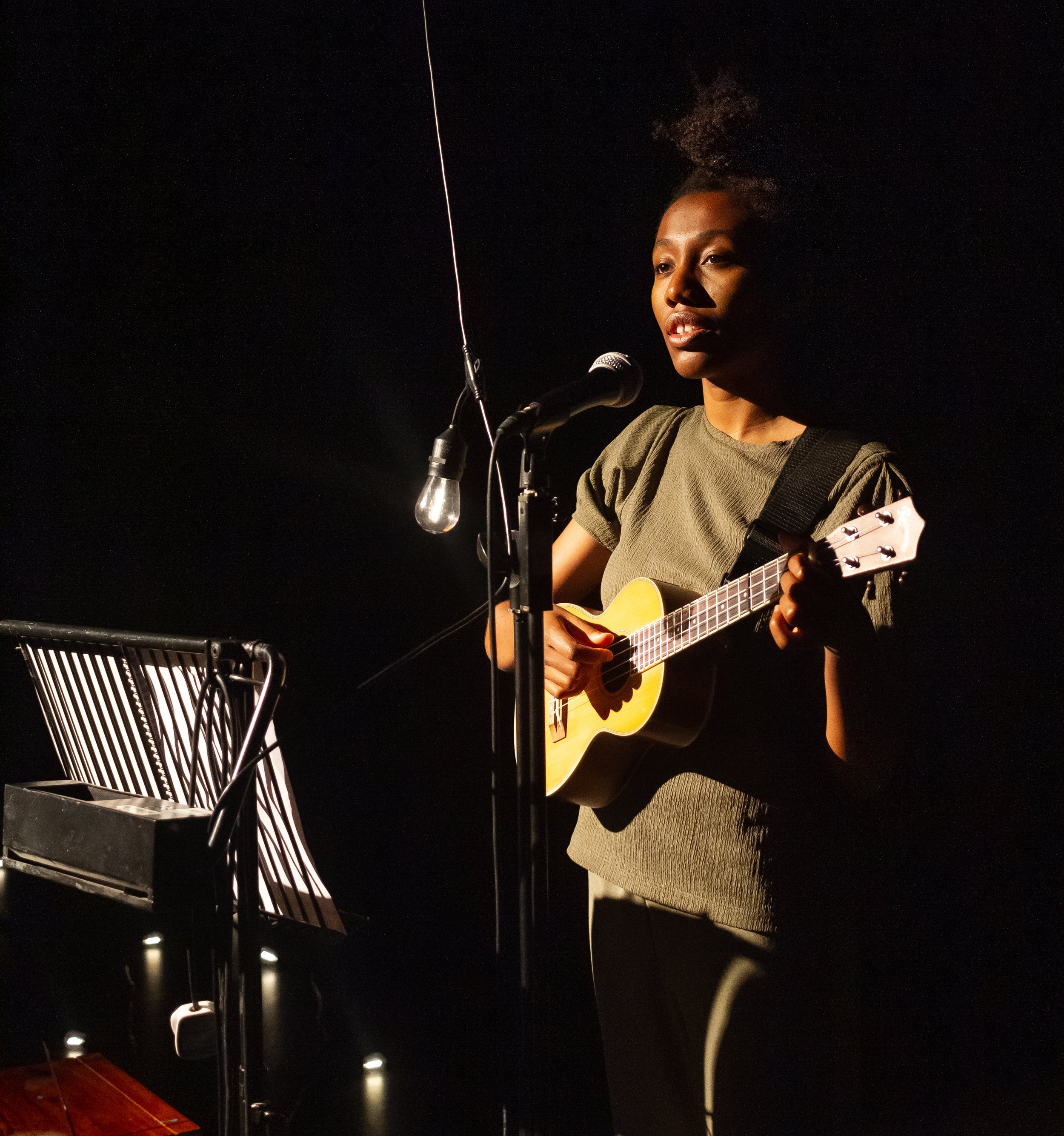  A Black woman is stood playing ukelele in front of a music stand and microphone a festoon bulb also hangs from the microphone stand  