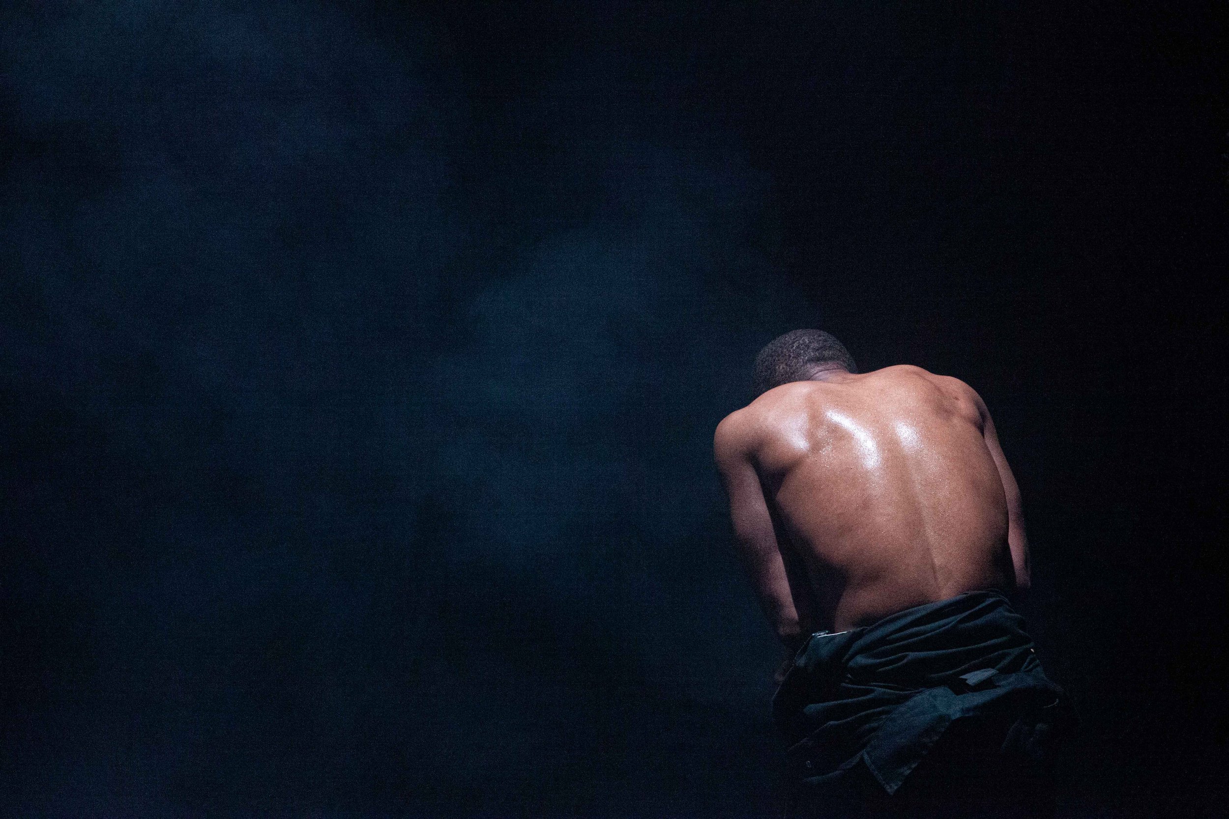  A shirtless man crouches in the corner of the dark image, his back to the viewer 