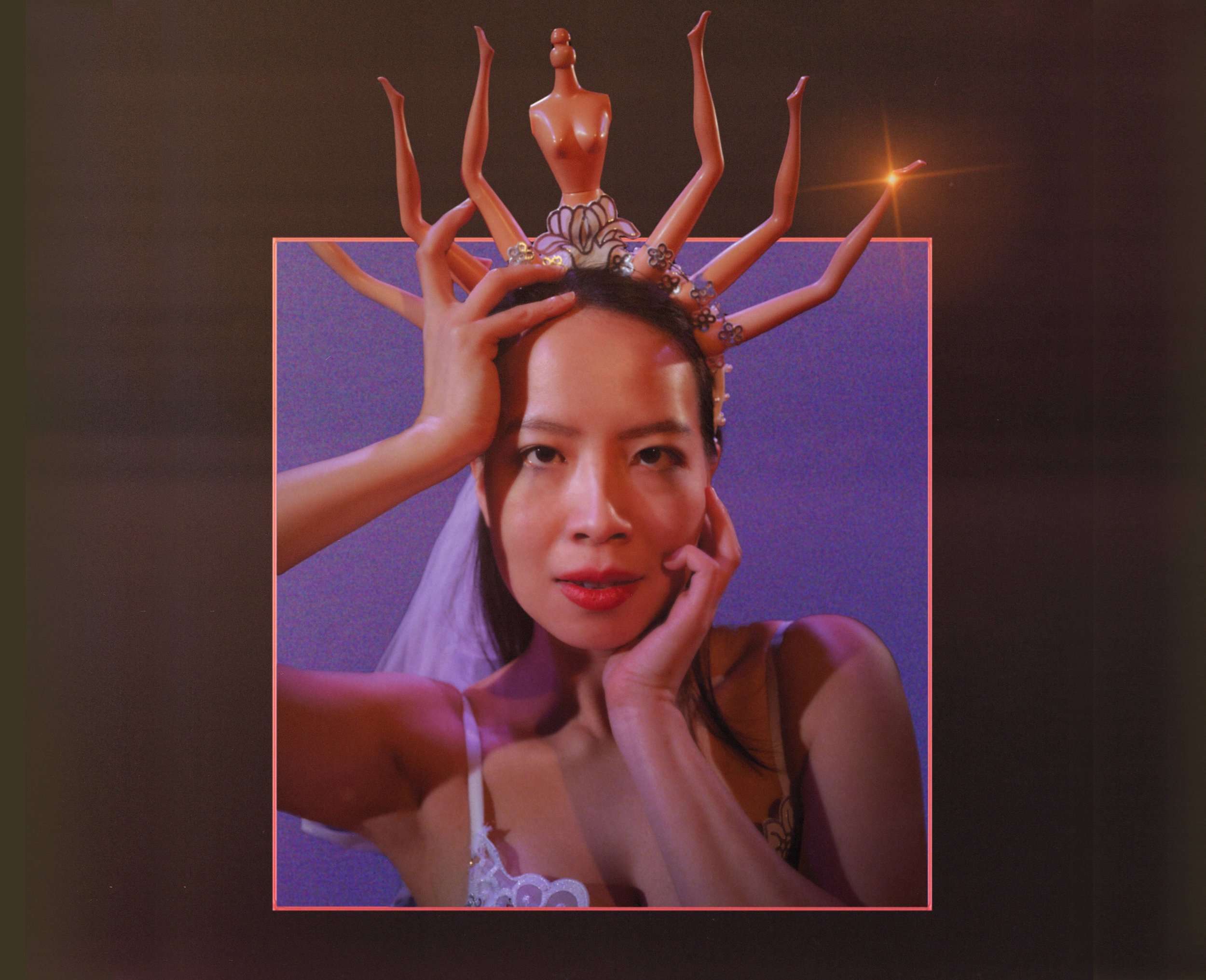  An East Asian woman strikes a post at the camera. She is wearing a white wedding train and a tiara made of Barbie Doll body parts 