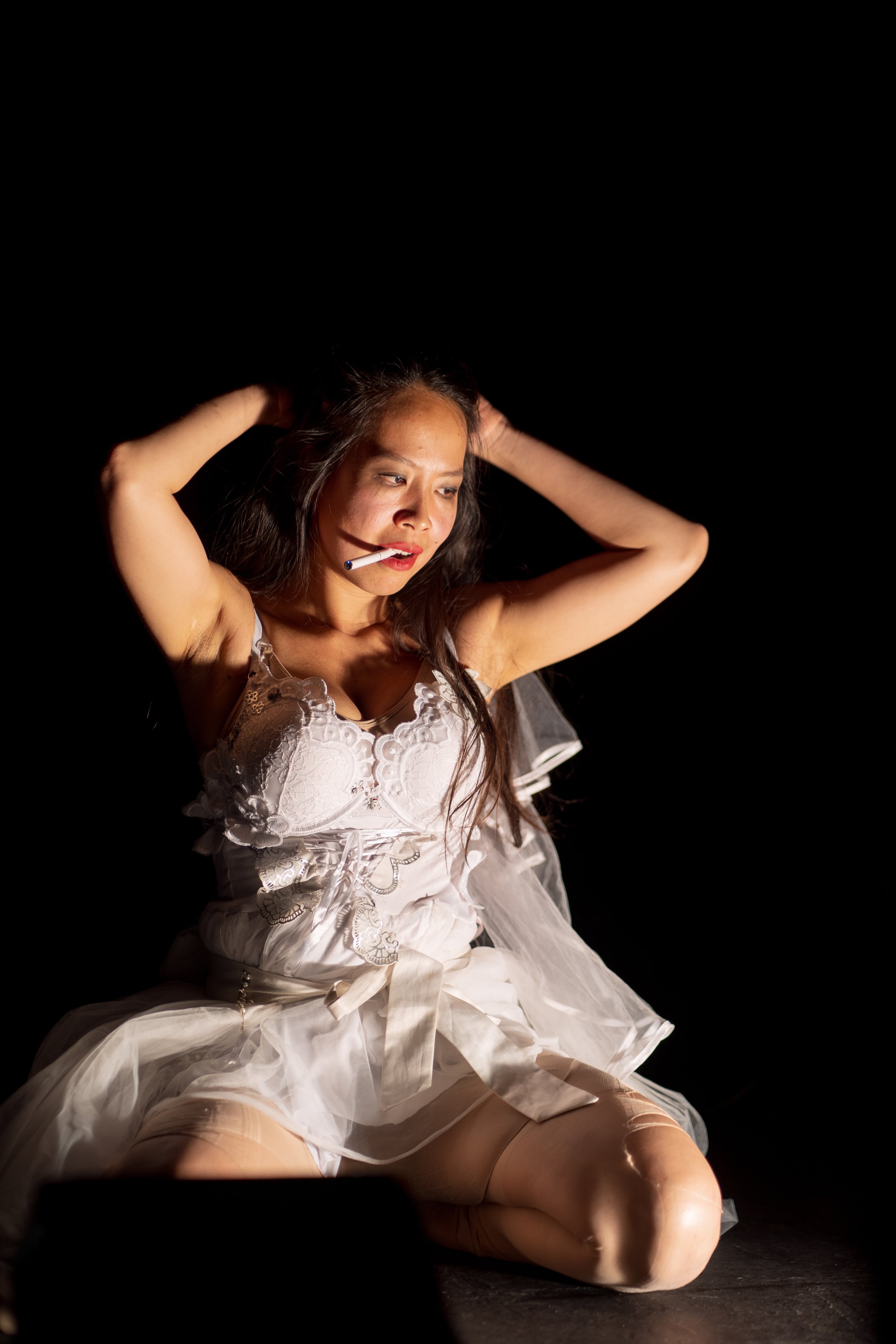  An East Asian woman in a wedding dress kneels on the floor. Her makeup is smudged and a cigarette dangles from her lips. 