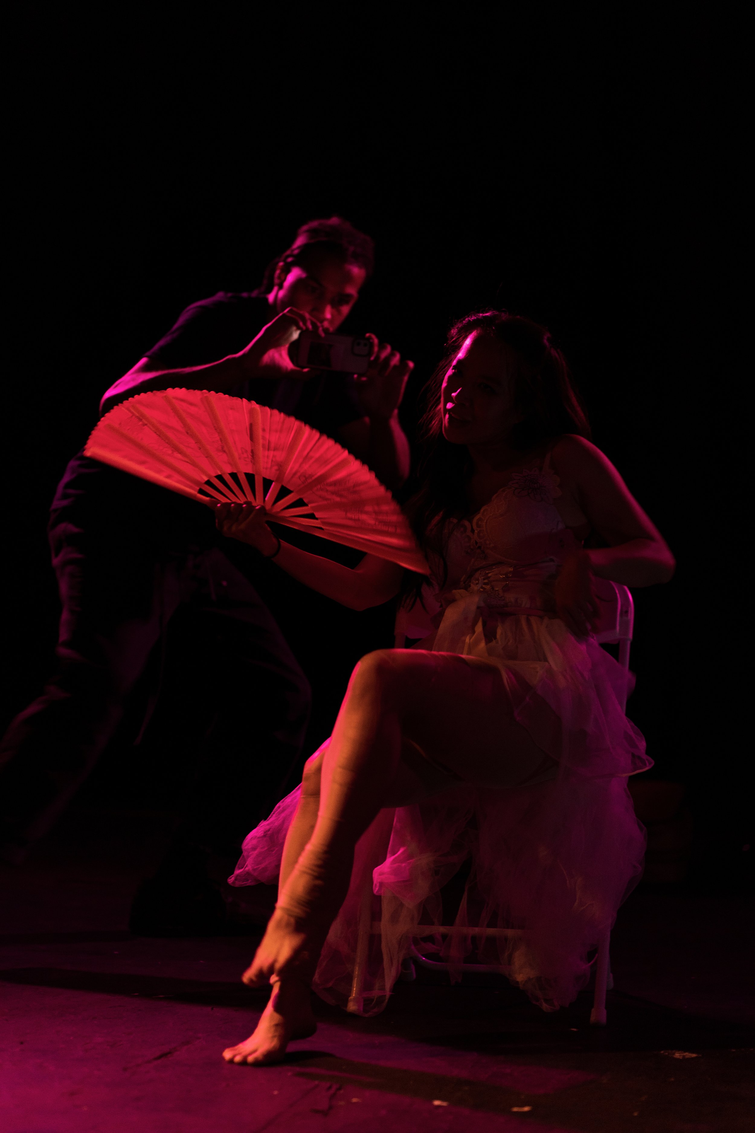  Bathed in a red light, a woman poses seductively on a chair whilst coquettishly holding a paper fan. A man in the background is taking a video of her on a phone. 