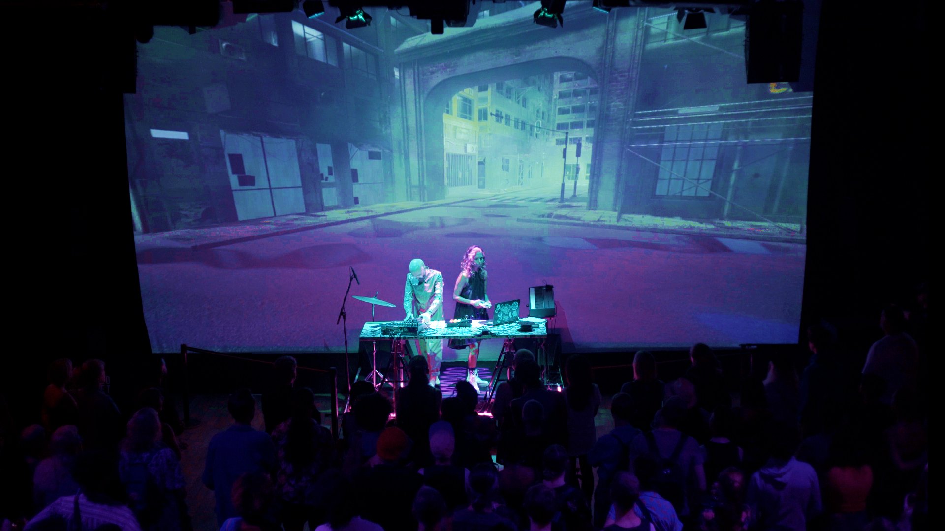  A woman with a games controller and a man with synth on stage playing music in front of giant projected image of a video game slum street 