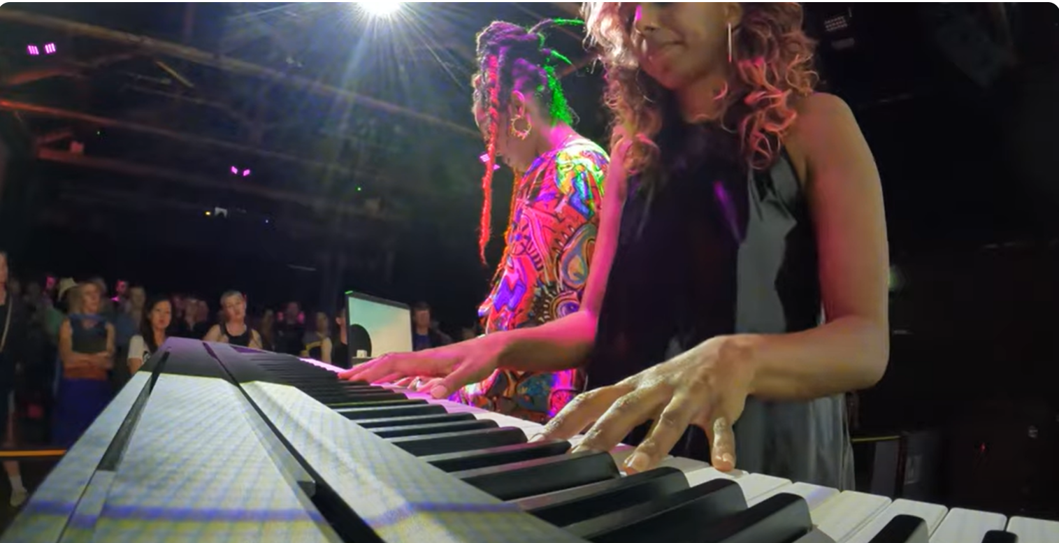  A close up of a woman playing a keyboard and another woman playing a video game live in front of a large audience 