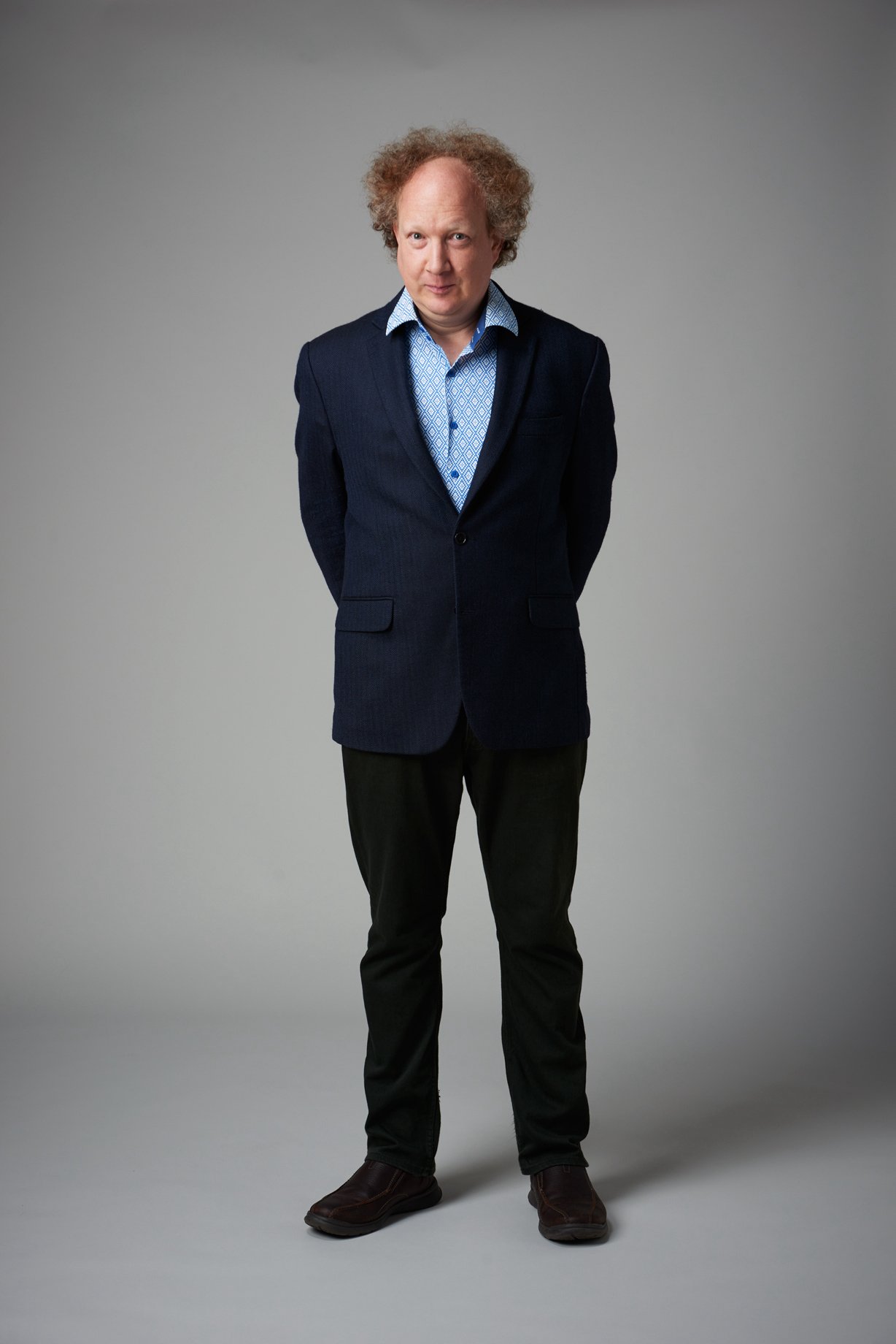  A full-length shot of Andy Zaltzman - a white, balding older man with curly brown hair. He is wearing a navy blue suit and standing with his arms behind his back whilst he smiled secretly at the camera. 