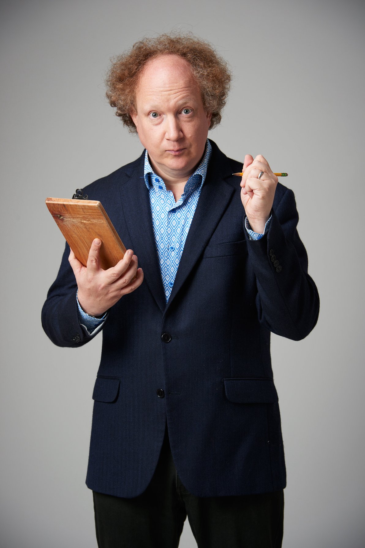  Andy Zaltzman - a white, balding older man with curly brown hair - looks quizically up from his clipboard and at the camera. 
