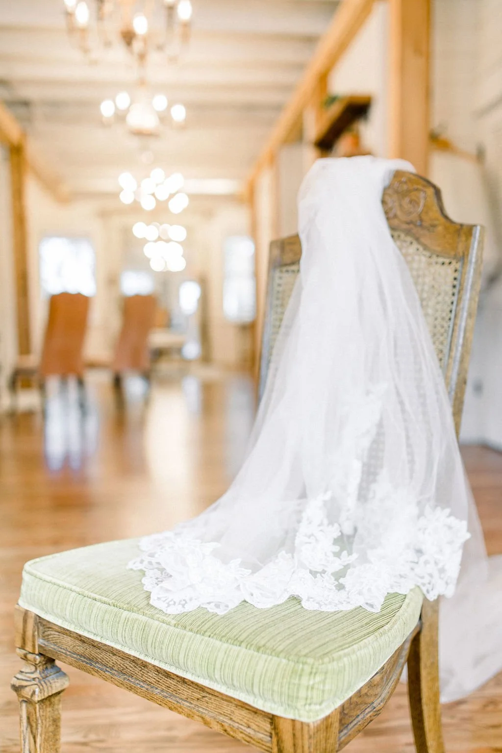 A wedding veil draped over the back of an antique wooden chair with a green upholstered seat, in a warmly lit room with chandelier and wooden beams.