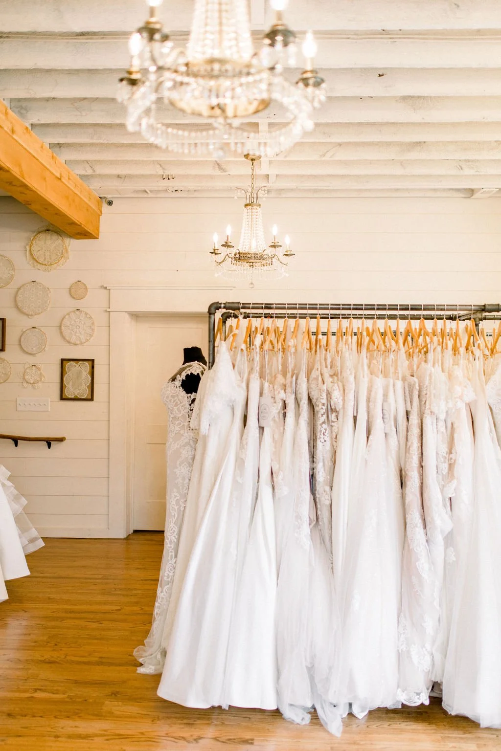 A boutique displays wedding dresses on a rack, with chandeliers hanging from the ceiling and circular woven art on the wall.