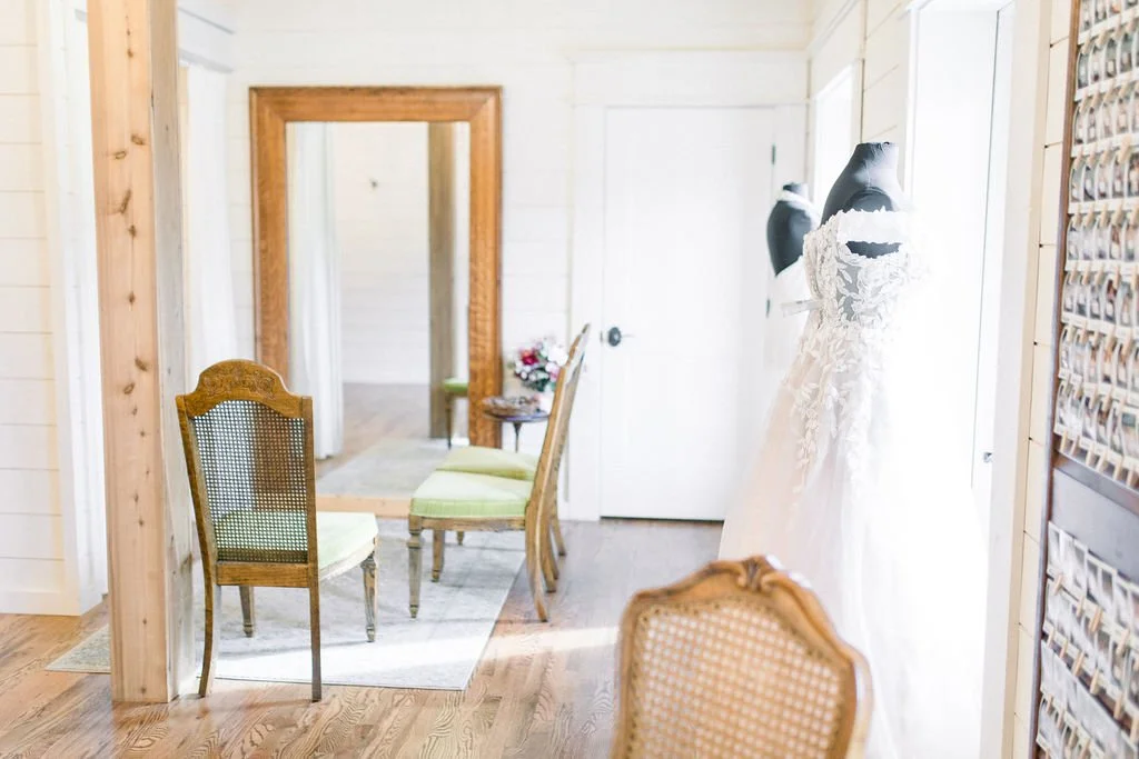A wedding dress on a mannequin in a bright room with wooden floors and white walls, a mirror, chairs, and a table with flowers.