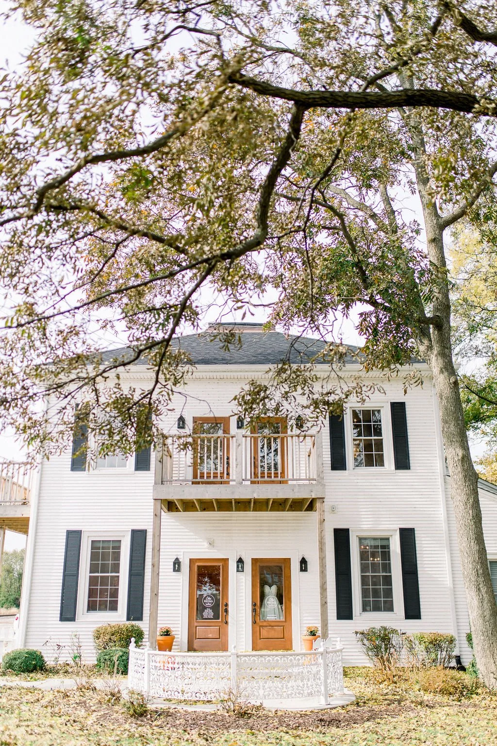 White two-story house with black shutters, two front doors with glass, and a small balcony, surrounded by trees and a white decorative fence.