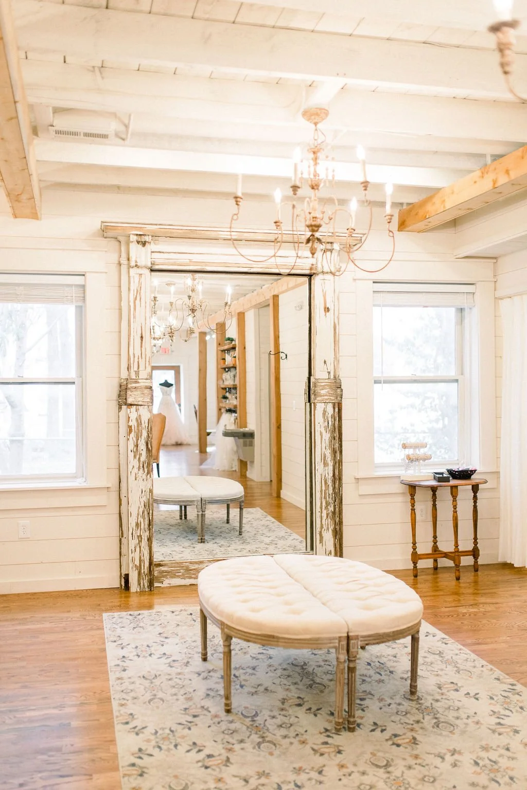 A bright, rustic interior room with white wooden walls and ceiling, hardwood floor, and large windows. A chandelier hangs from the ceiling. In the foreground, there is a tufted cream-colored bench on a patterned rug. A small wooden side table with de