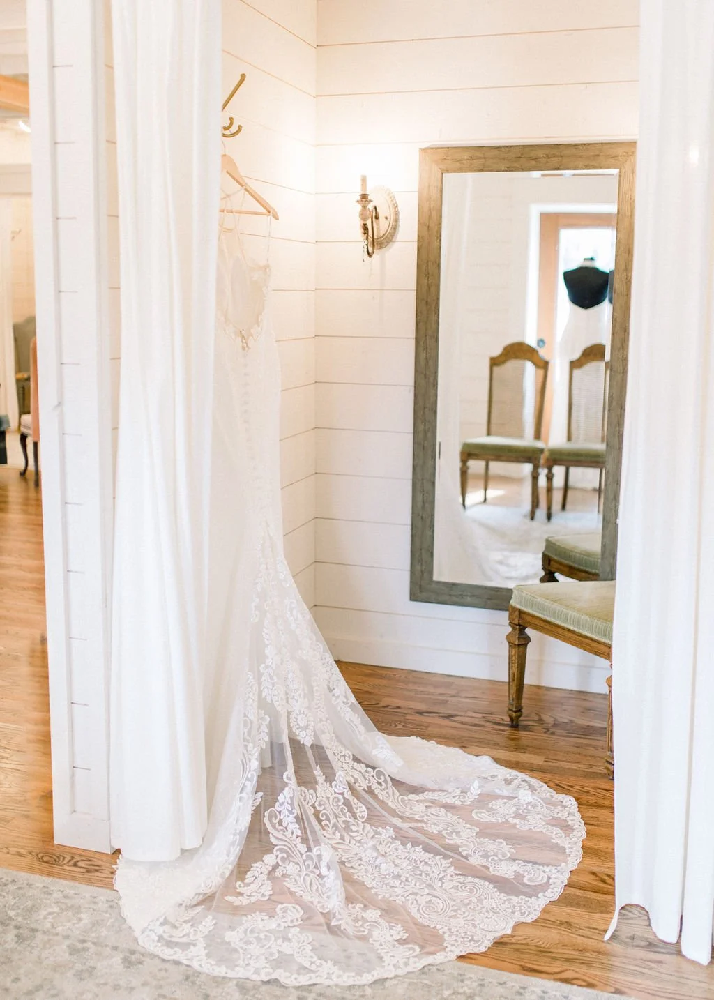 A white wedding dress hanging on a hook, reflected in a large wooden-framed mirror in a rustic, light-colored wood-paneled room with chairs and a small black dress form visible.