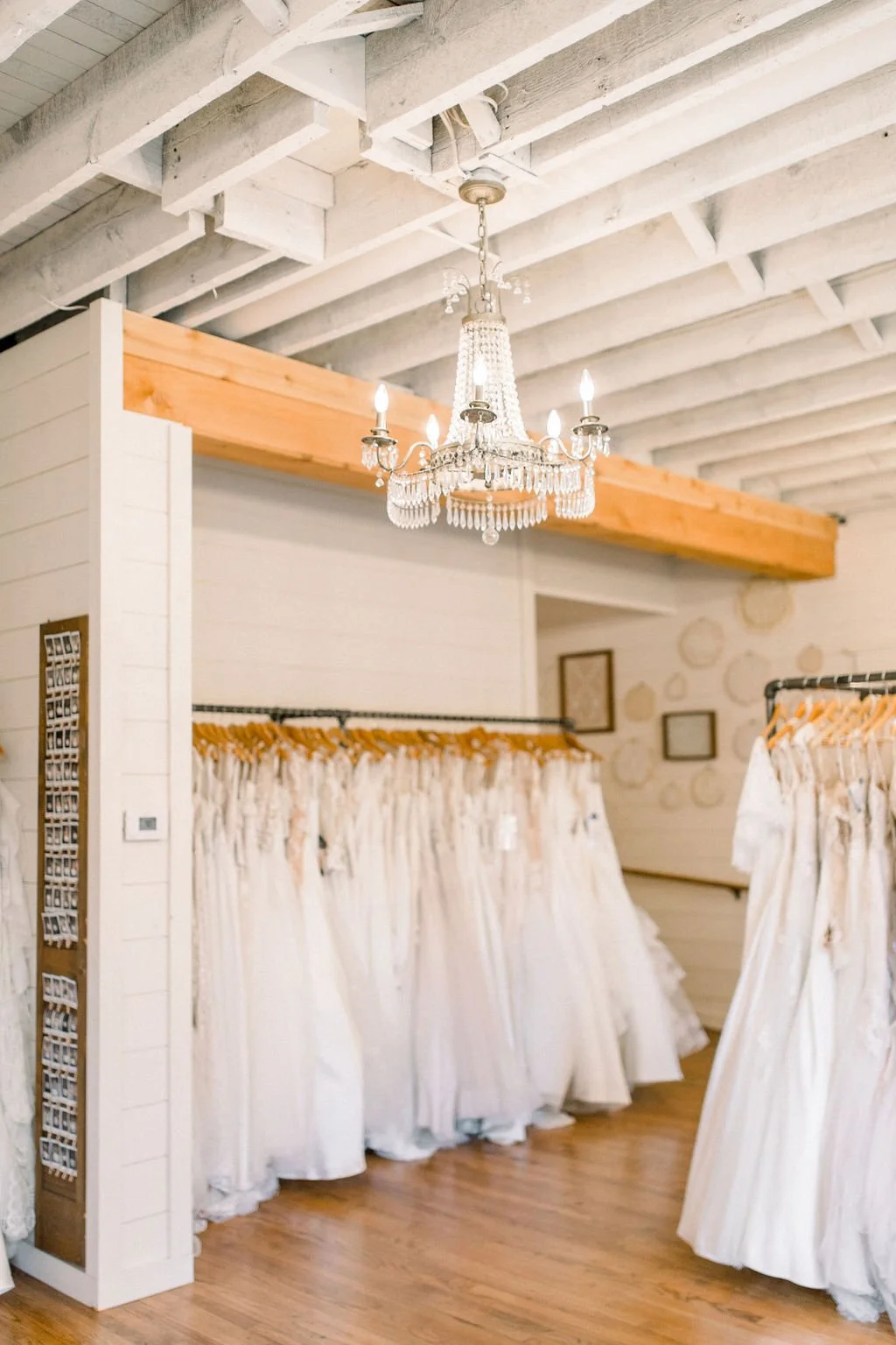 Interior of a bridal shop with wedding dresses on racks, a chandelier hanging from the ceiling, and a wooden beam.