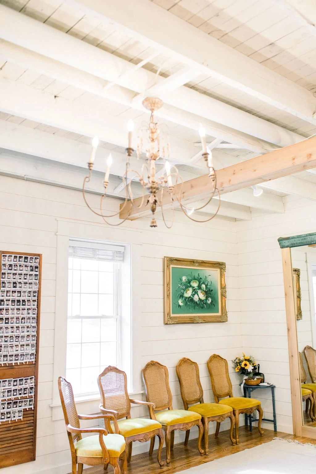 A vintage chandelier hanging from a white painted wooden ceiling in a bright room with white shiplap walls. There are five antique-style chairs with yellow velvet cushions lined up against the wall, a small table with a flower arrangement, a framed f