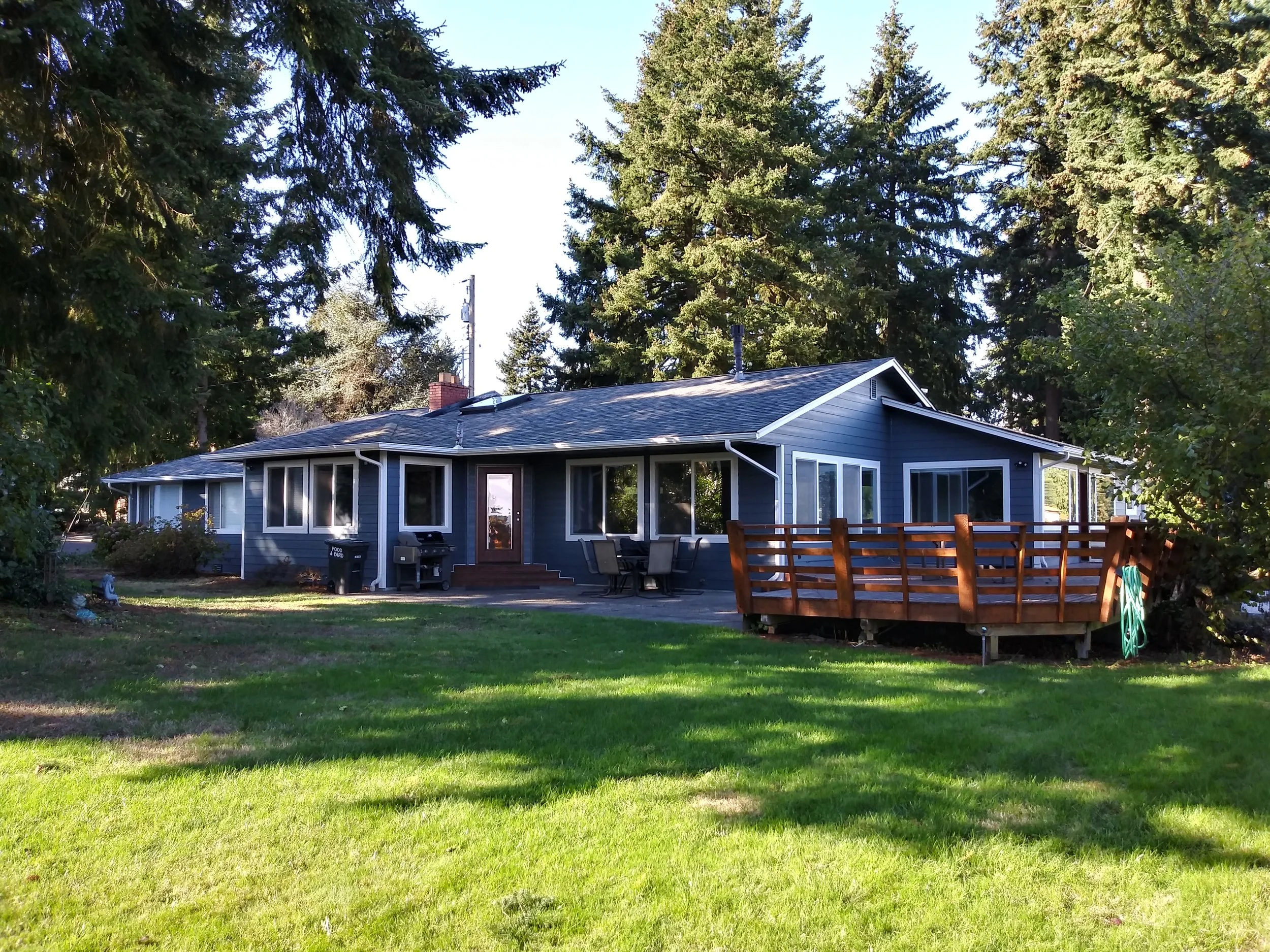 Single-story home with dark blue fiber cement siding, white-framed windows, and a custom wood deck, surrounded by evergreen trees in a Pacific Northwest setting.