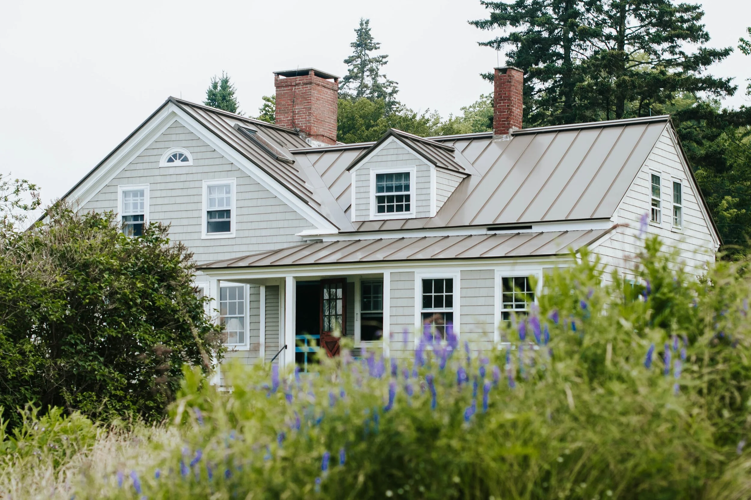 A charming two-story farmhouse-style home with light gray shingle siding and a standing seam metal roof in a bronze color. The house features white trim around the windows, two prominent red brick chimneys, a front porch with a Dutch door, and is surrounded by lush greenery and blooming purple wildflowers in the foreground.