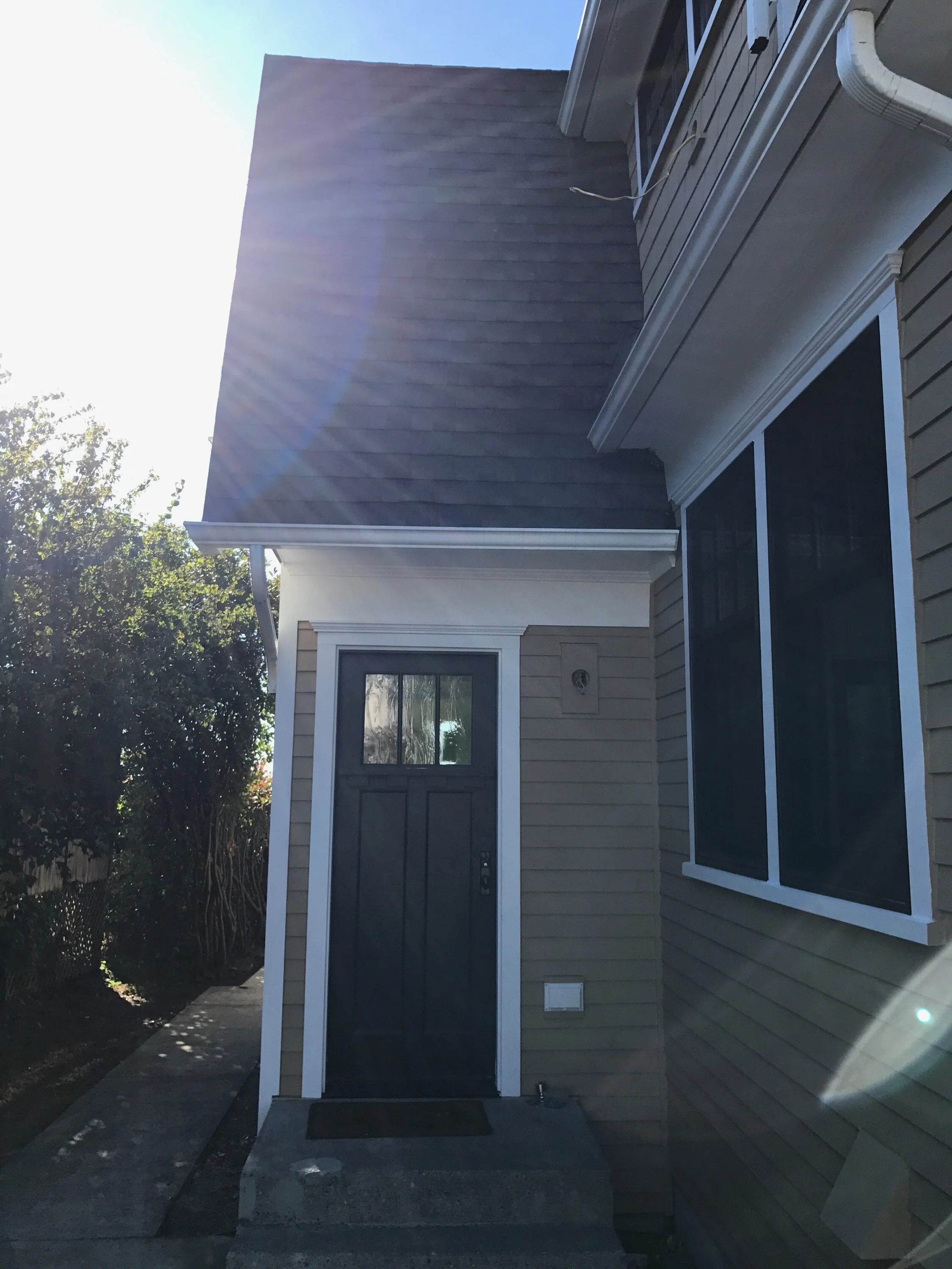 Side entrance of a two-story house with tan horizontal siding, white trim, and a black front door with window panes. A small concrete stoop leads to the door, and a narrow pathway runs alongside the house under bright sunlight with visible lens flare.
