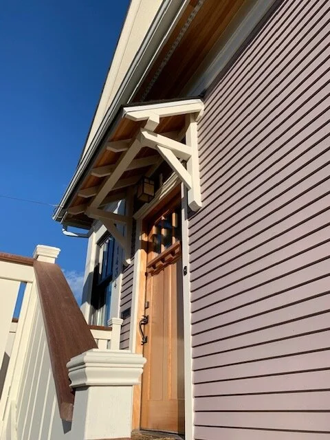 Close-up view of a newly installed wood front door with decorative overhang and detailed trim, paired with fresh lap siding and a custom porch railing.