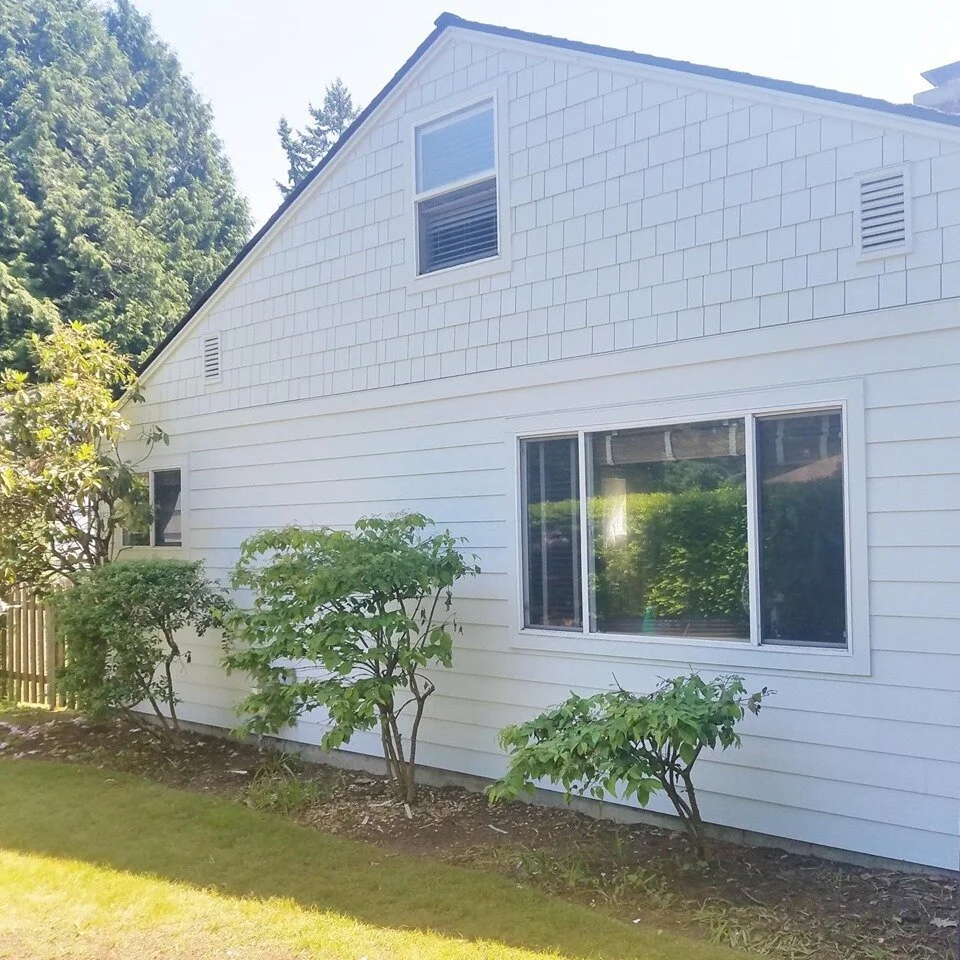 Side view of a single-story house with light-colored horizontal lap siding on the lower half and white shingle siding on the gable. The house features several white-trimmed windows and a few small bushes and trees planted along a narrow garden bed next to a green lawn.