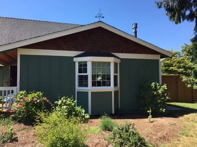 Single-story house with green vertical board-and-batten siding and brown shingle gable. A white-trimmed bay window projects from the front, surrounded by a small landscaped garden with flowering plants and shrubs under a clear blue sky.