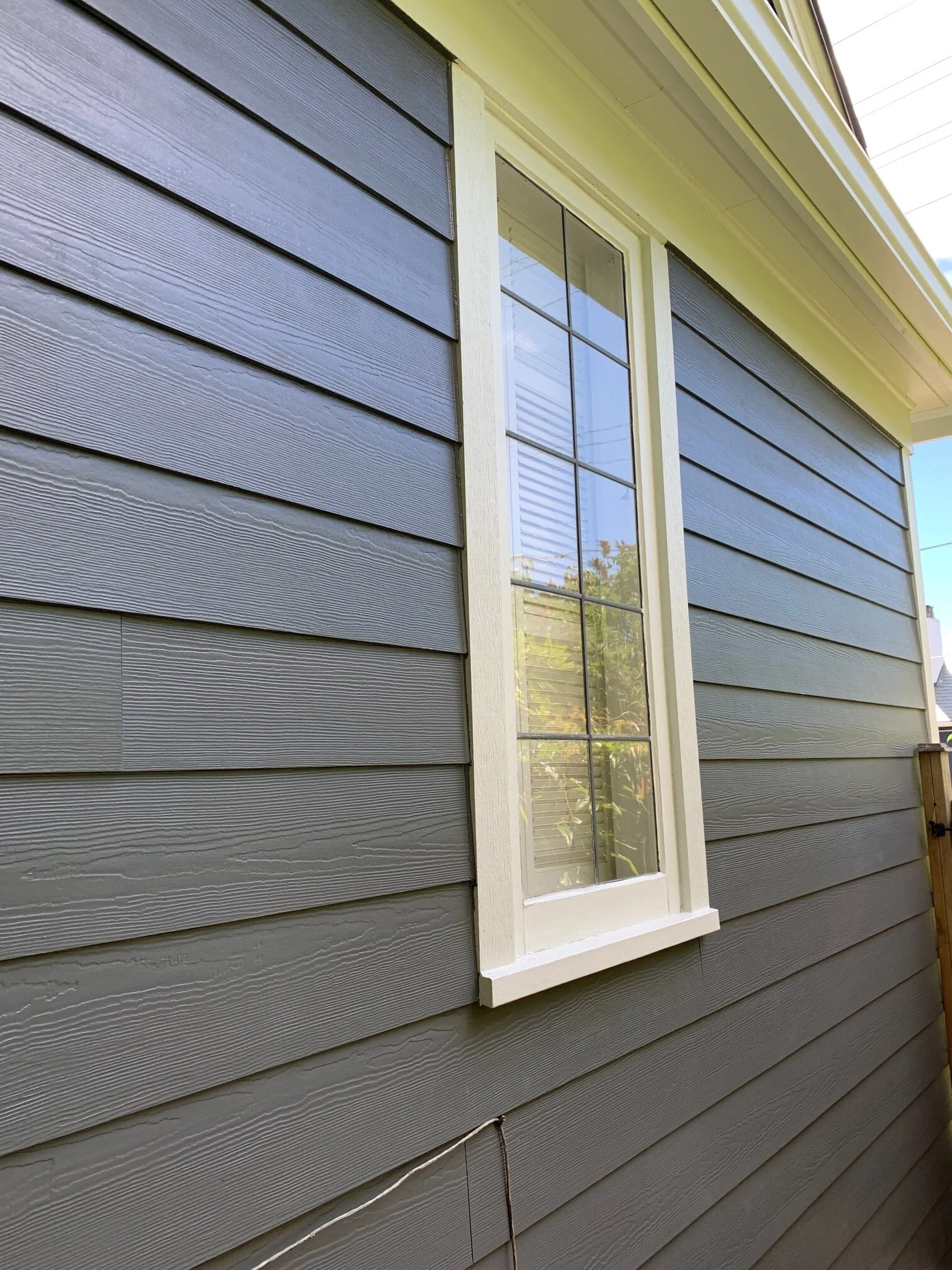 Close-up view of a house exterior with horizontal fiber cement siding painted in a dark gray color. The image shows a narrow vertical window with white trim and decorative grid lines, reflecting the surrounding yard and sky.
