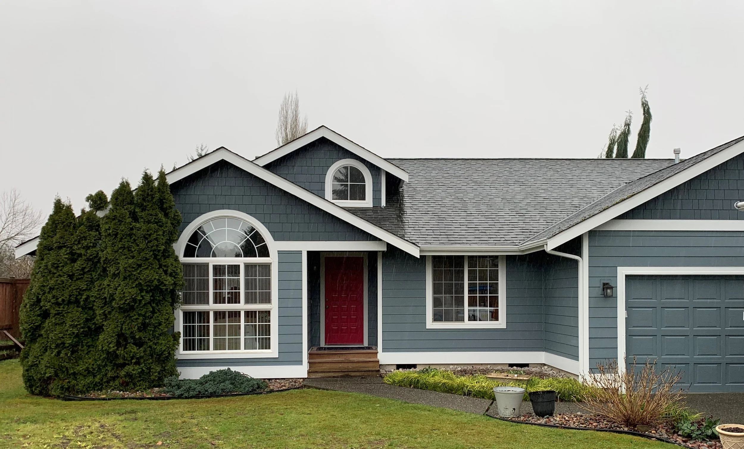 Single-story home with dark blue lap and shake siding, white trim, and arched windows, featuring a red front door and gray shingle roof—highlighting a full exterior siding renovation
