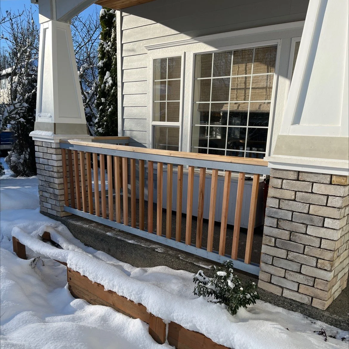 Front porch with natural wood railing, white lap siding, and brick column bases, pictured in a snowy winter setting—highlighting craftsmanship in siding and exterior trim work