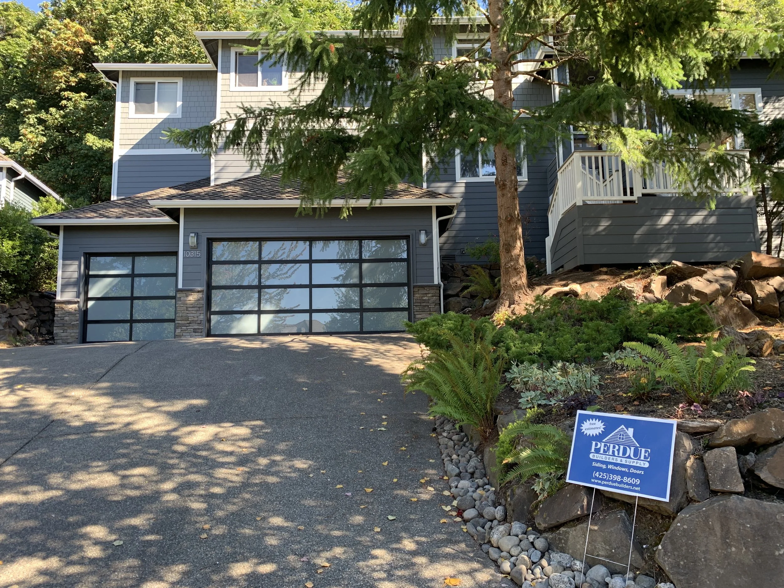 Modern two-story home with gray siding, large glass garage doors, and stone trim, featuring Perdue Builders' sign for expert siding, window, and door replacement services.