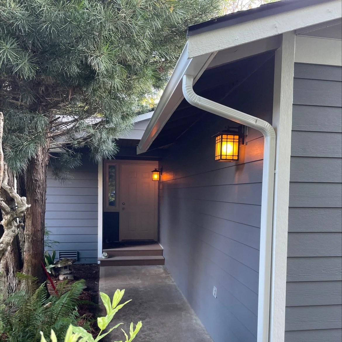 Cozy covered entryway with gray horizontal lap siding, warm exterior lighting, and white trim, surrounded by lush greenery—highlighting a welcoming home exterior renovation.