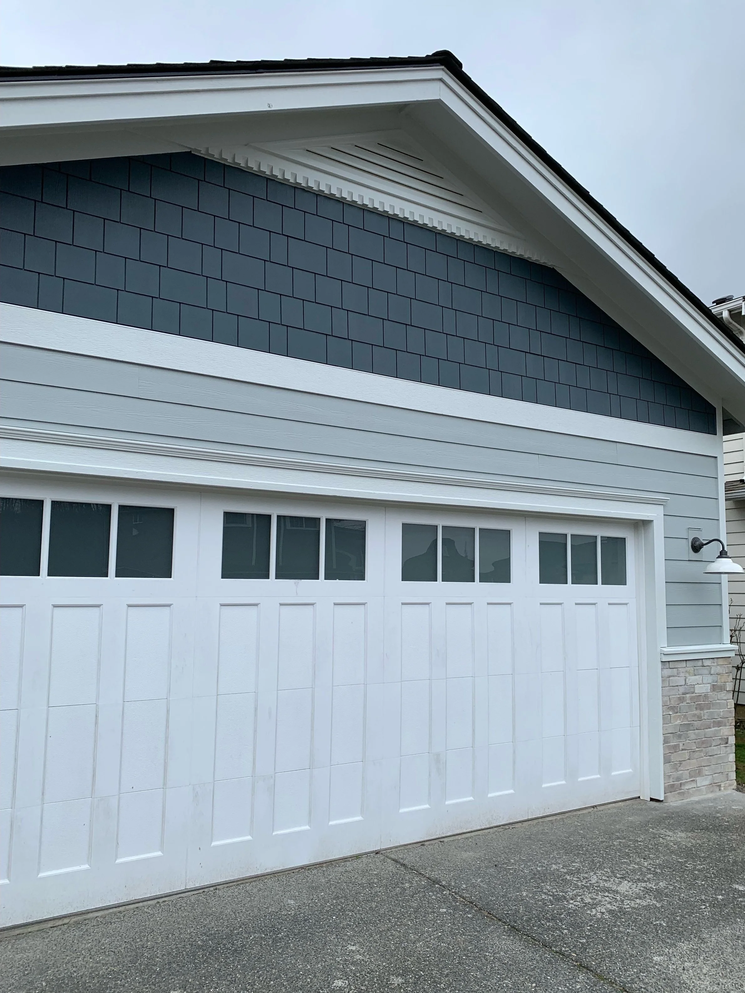 A house garage exterior featuring a white paneled garage door with upper windows, light gray horizontal siding, and a gable clad in dark blue square shingles. The trim is painted white, and there is a small white outdoor light fixture mounted on the right side above a section of light-colored brick veneer.