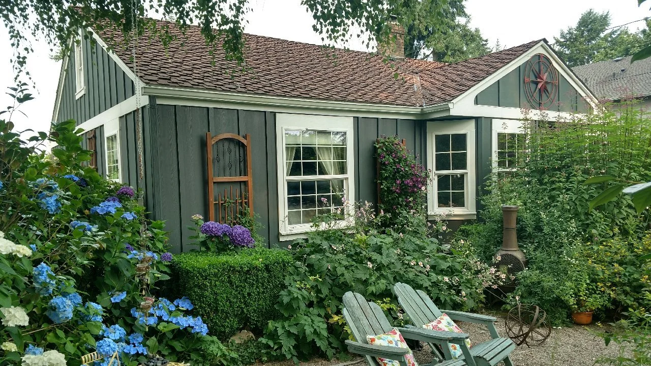 Charming cottage-style house with dark green vertical siding, white trim, and a reddish-brown shingle roof, surrounded by lush gardens filled with colorful flowers, shrubs, and vines. Two Adirondack chairs with floral pillows sit in a small gravel patio area, alongside a rustic outdoor fireplace.