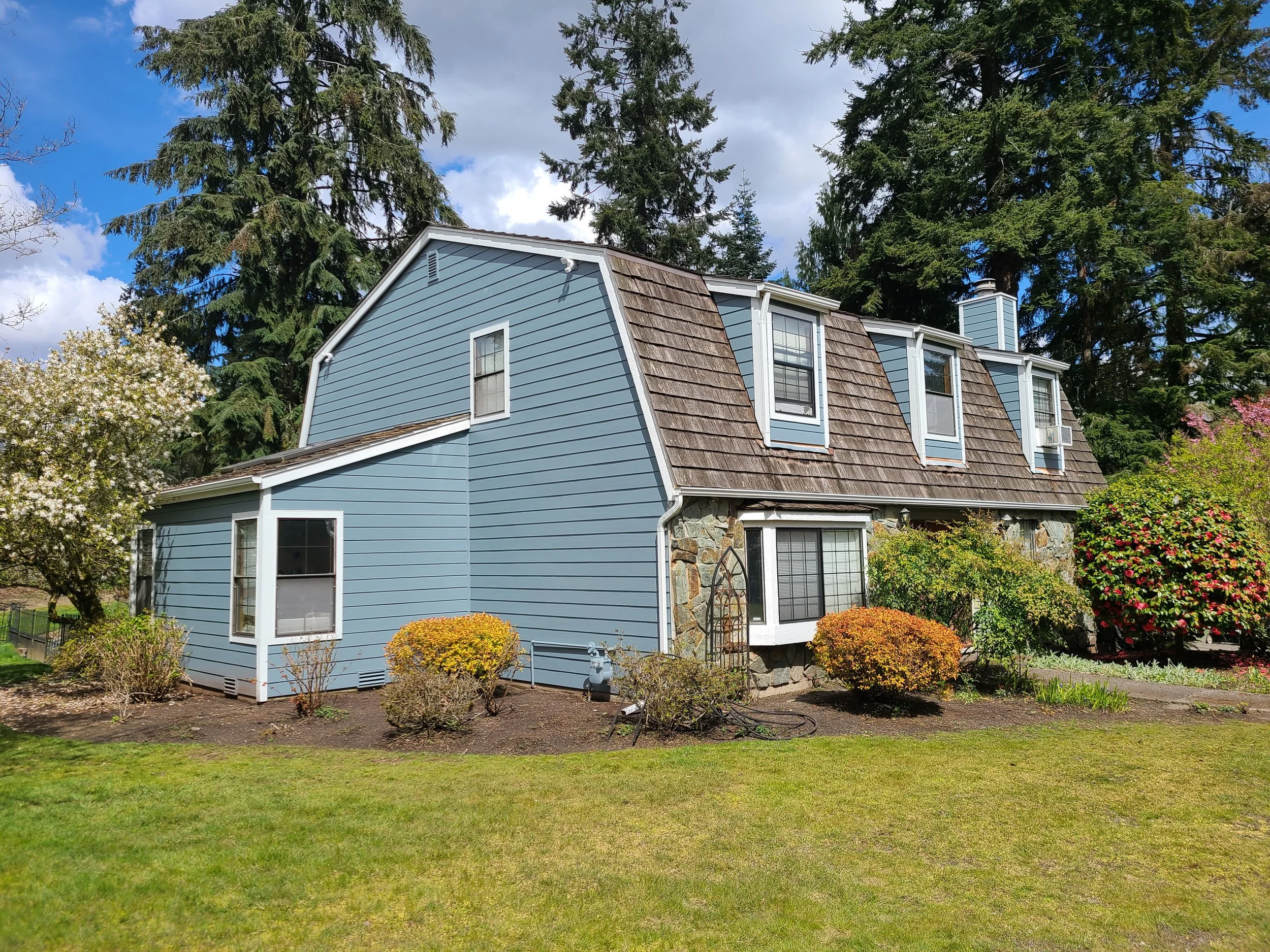 Two-story home with blue horizontal lap siding, cedar shake accents, and stone veneer detailing, surrounded by lush landscaping—highlighting a custom exterior renovation project.