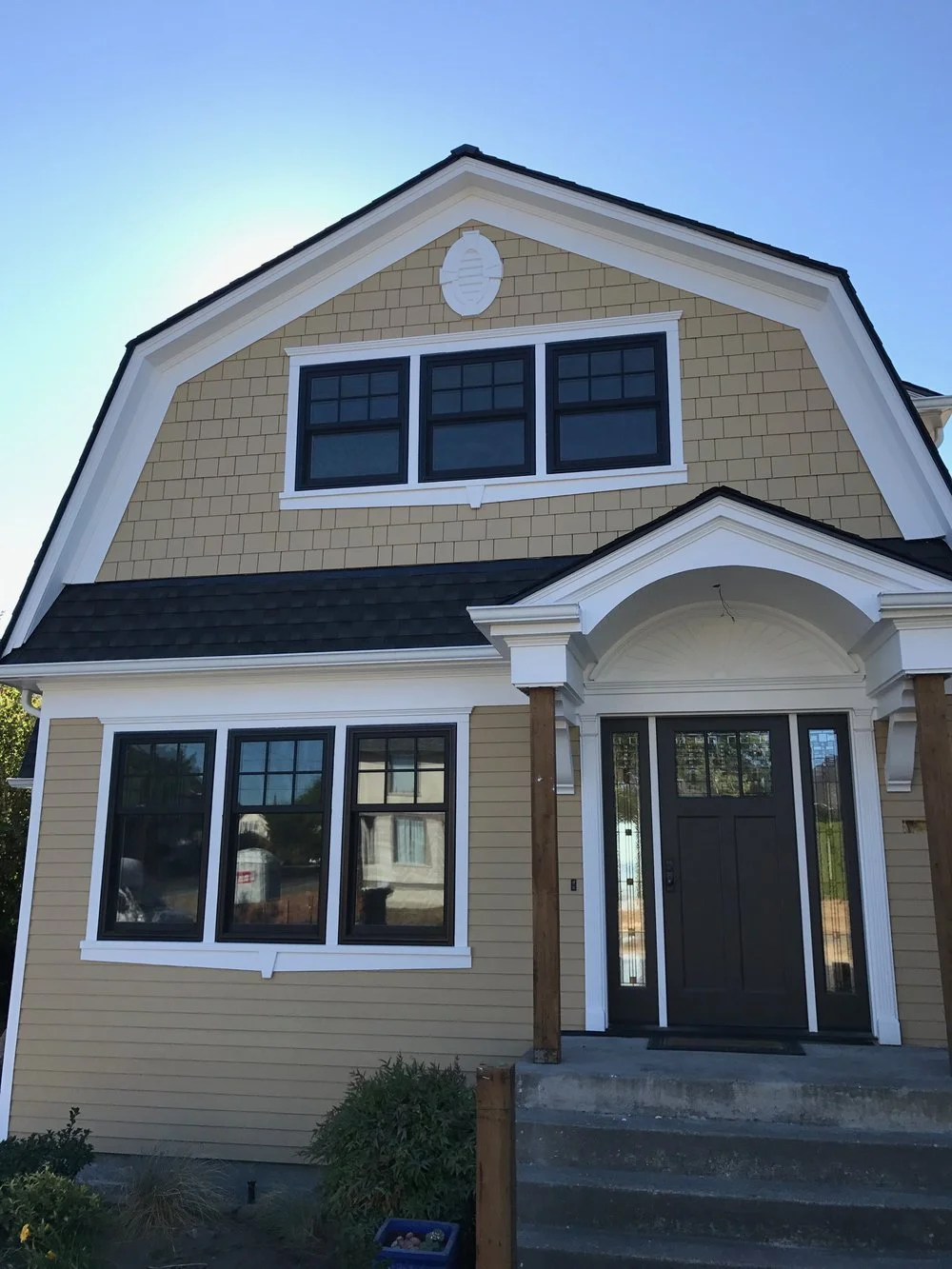 A front view of a two-story house with a gambrel roof. The upper section features tan-colored shake siding with black-trimmed rectangular windows. The lower section has horizontal tan siding with black window trim and white framing. The entryway includes a dark brown front door with glass panes, white columns, and an arched pediment above.