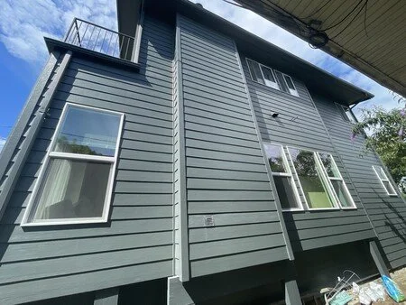 Upward angled view of a modern two-story house with dark green horizontal siding and white-trimmed windows. The house features a small upper balcony and multiple windows on both floors, with shadows cast from the roofline and surrounding structures under a partly cloudy sky.