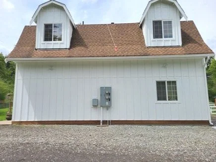 White barn-style home with vertical board and batten siding and brown shingle roof, showcasing fresh exterior siding installation with updated windows.