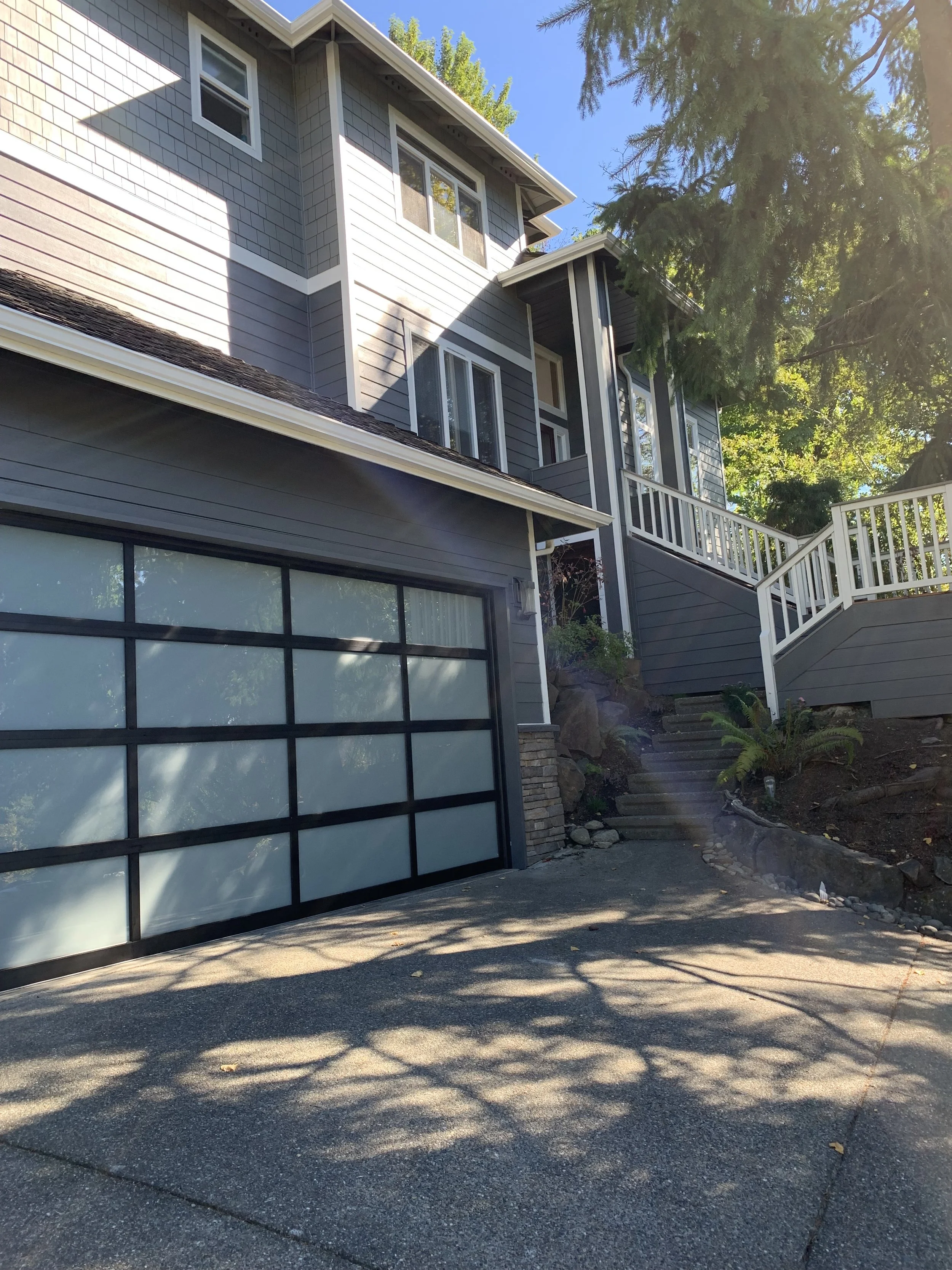Two-story modern house with gray horizontal and shingle siding, white trim, and a large glass-paneled garage door. A concrete driveway leads up to the garage, with a side staircase ascending through landscaped rocks to a white-railed porch entrance. Sunlight filters through nearby trees, casting shadows on the driveway.