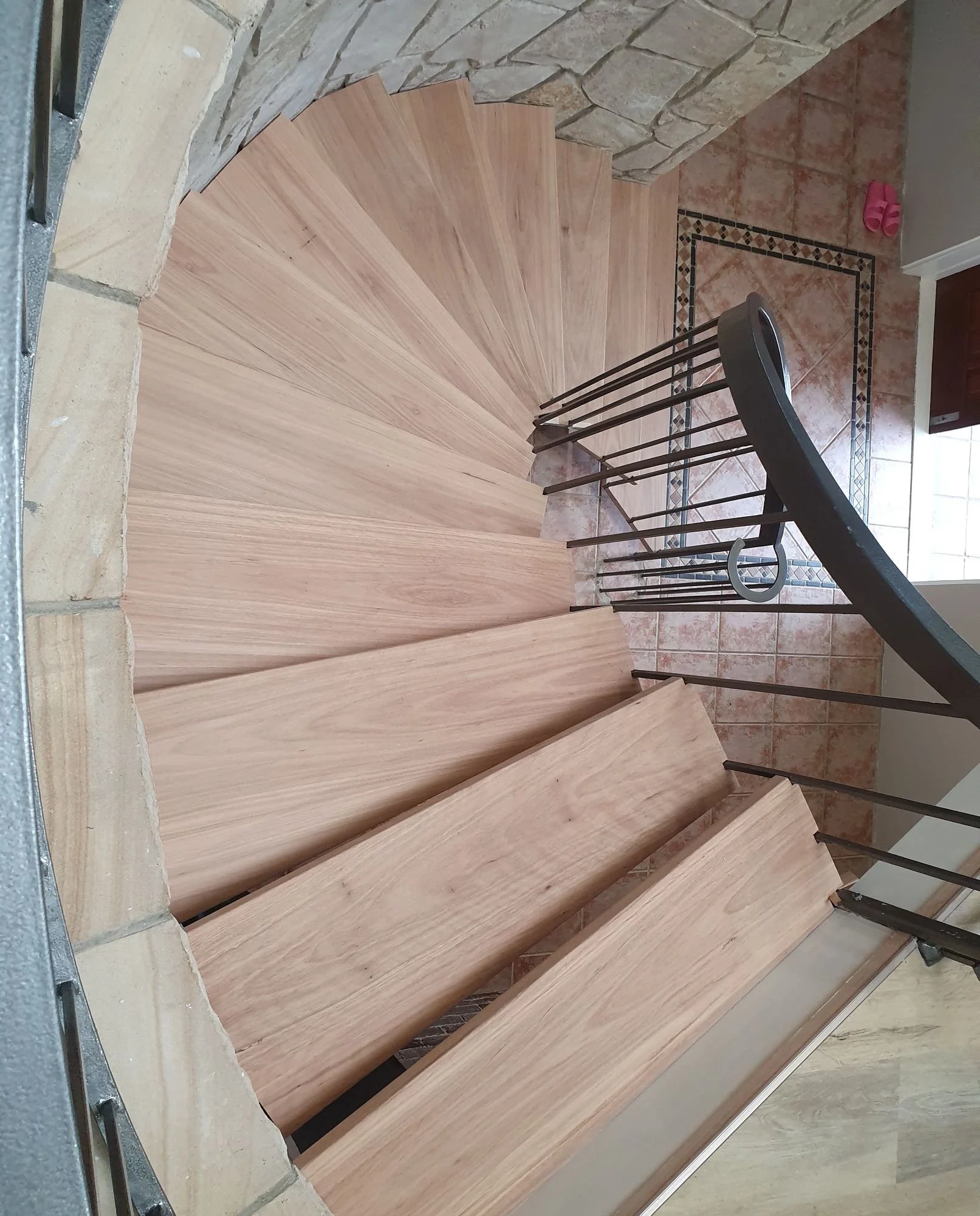  Looking down a timber spiral staircase curving against a sandstone wall, with a black steel handrail 