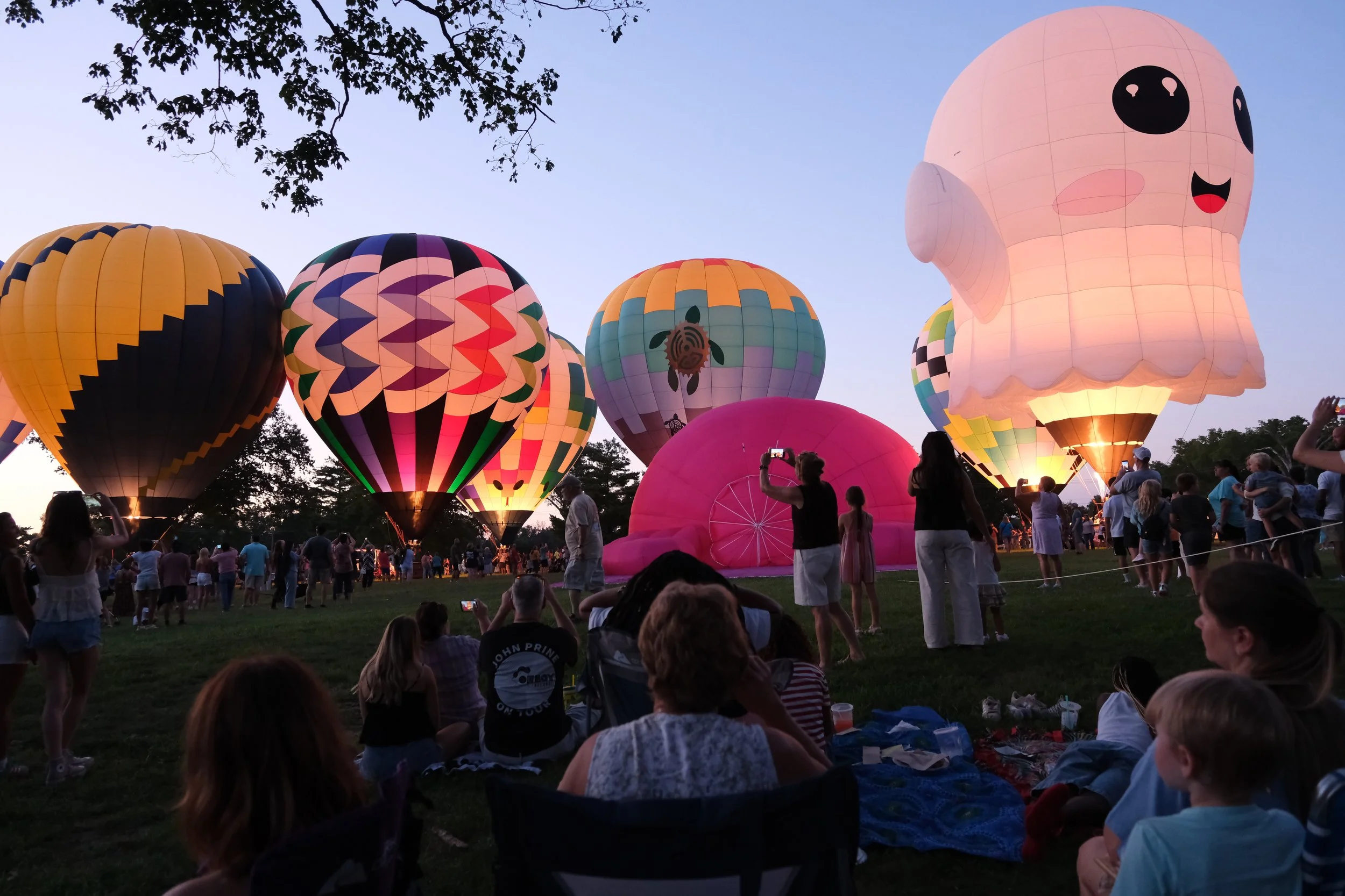 A group of colorful hot air balloons with various patterns, including a large pink balloon shaped like a cute cartoon character with big eyes and a smiling face, being prepared for flight at sunset with many people gathered around.