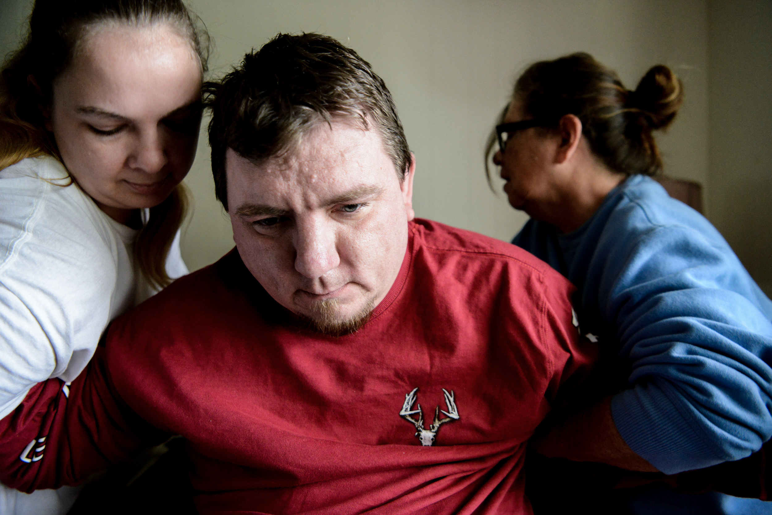  Adam waits for Chery and his medical aid to finish dressing him during their morning routine. It takes two people to get Adam ready every day, including changing his waste bags and moving him from the bed to a motorized wheel chair. 