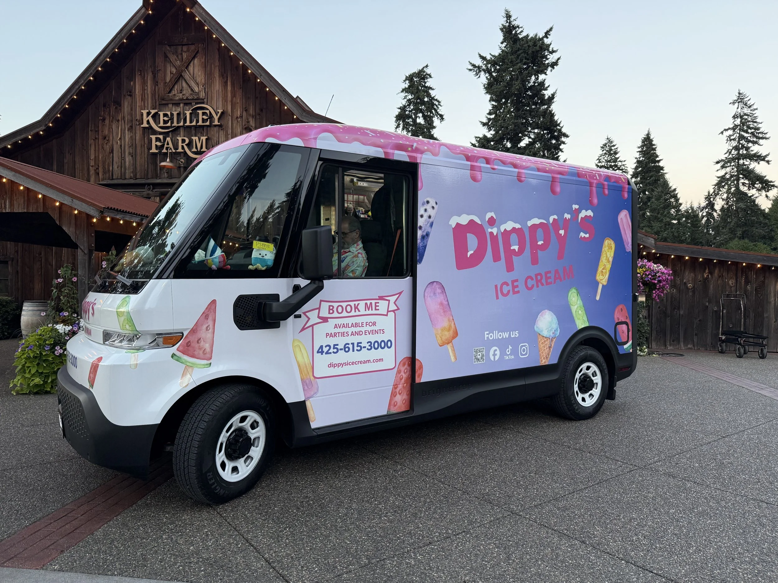 Ice cream truck with pink, blue, and yellow ice cream graphics parked in front of a rustic wooden building with a sign that reads 'Kelley Farm.' Flowers and trees are visible around the area.