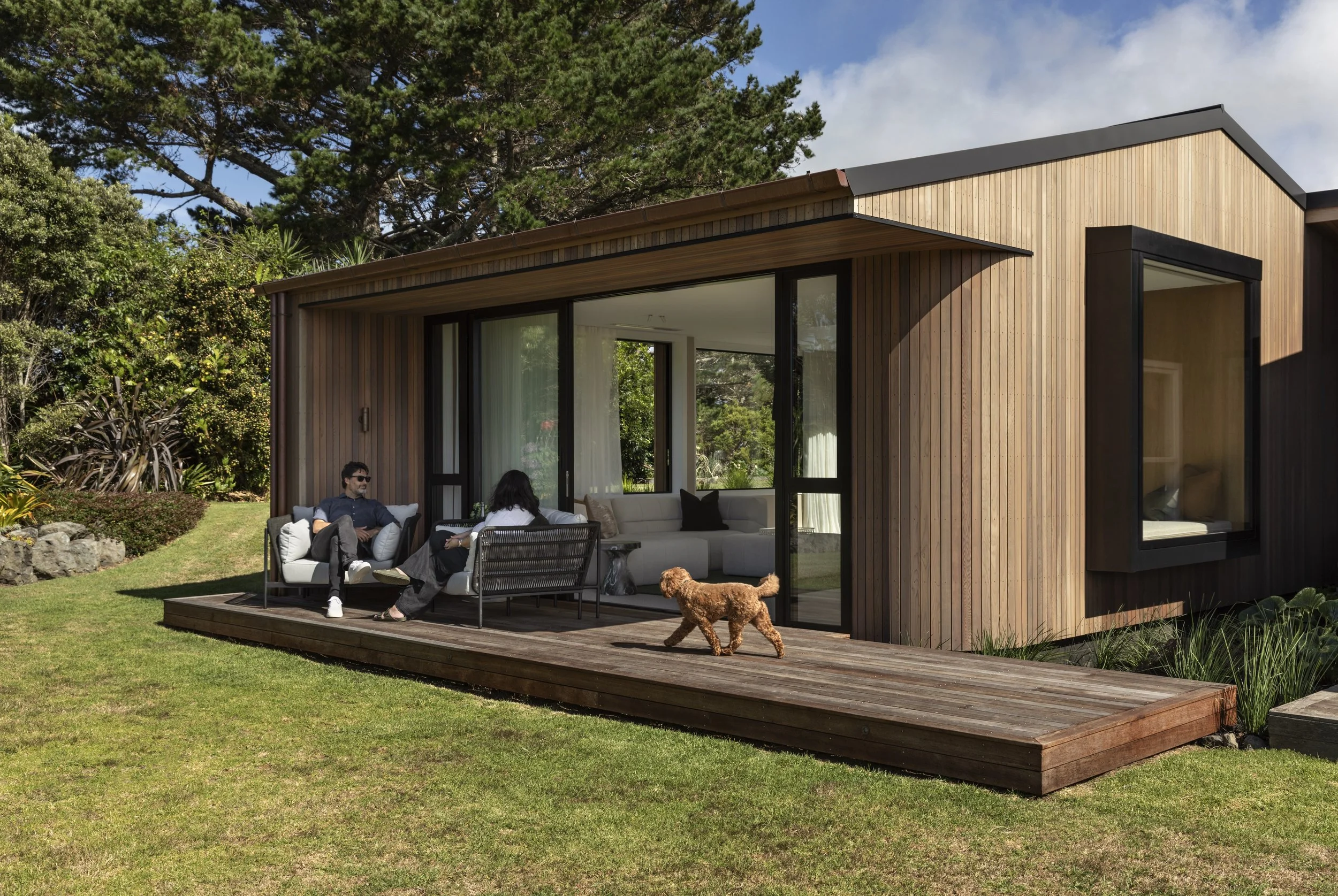 Modern wooden house with large sliding glass doors, outdoor patio with two people sitting and talking, dog walking on the wooden deck, surrounded by green grass and trees.