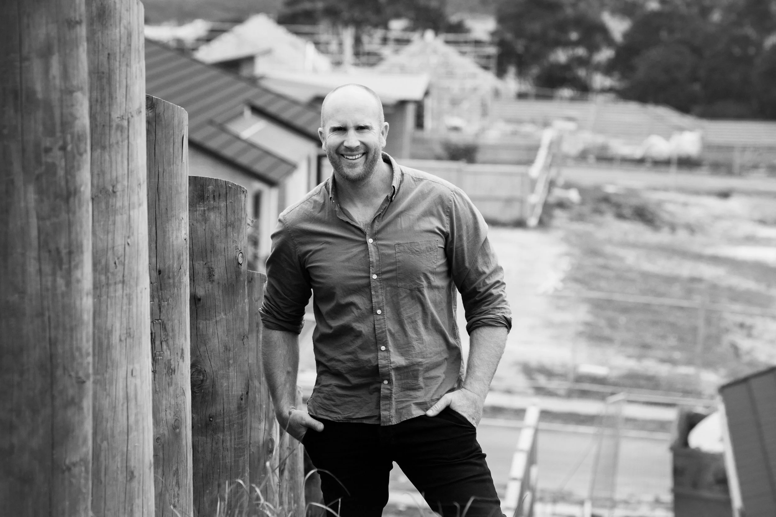 A smiling man stands outdoors next to a wooden fence, with a rural landscape behind him.