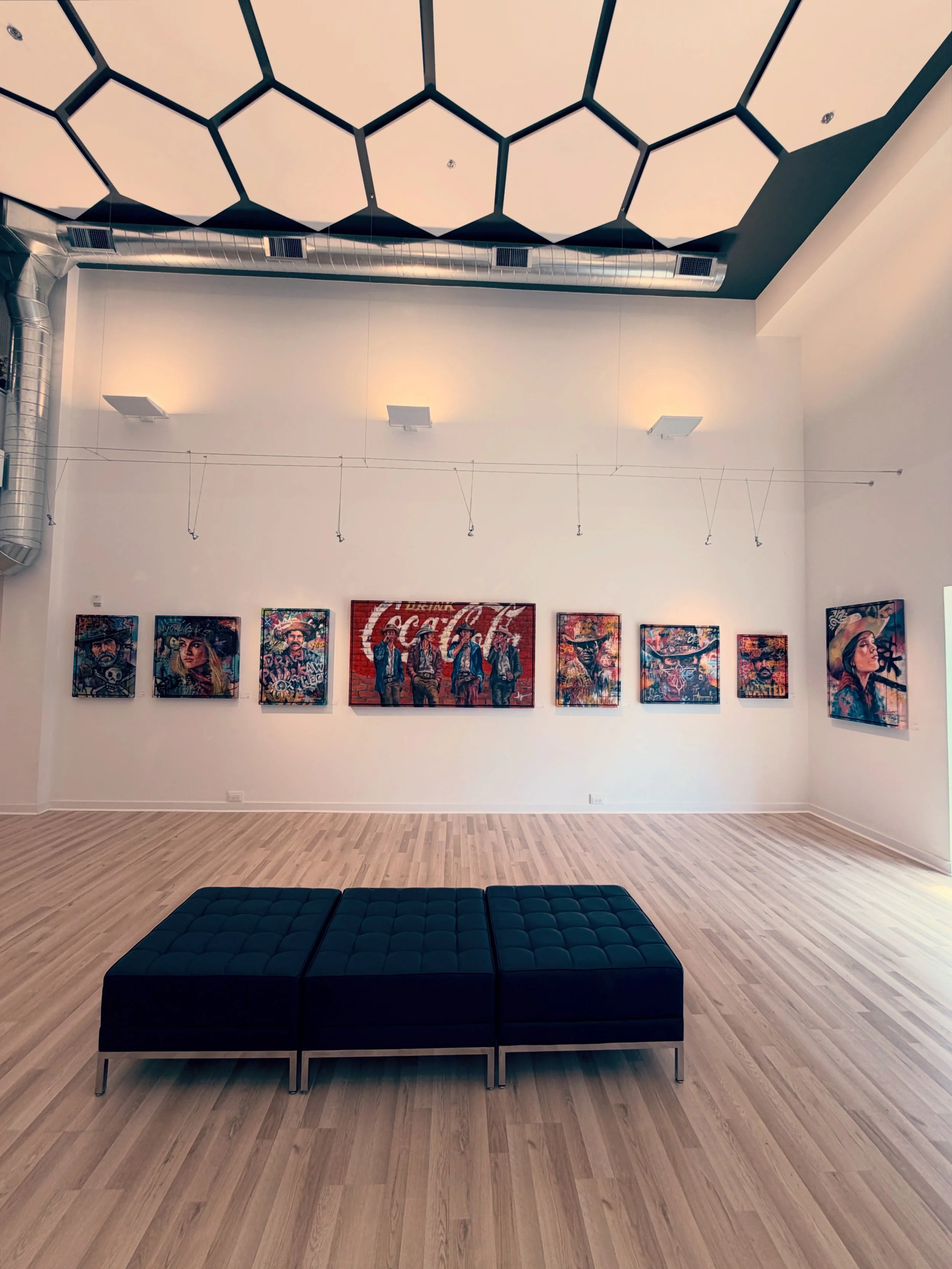 Art gallery with framed artwork on the wall including a central piece with the Coca-Cola logo and surrounding images of people, a black cushioned ottoman in the foreground, light wooden flooring, and a plain white wall with ceiling lights.