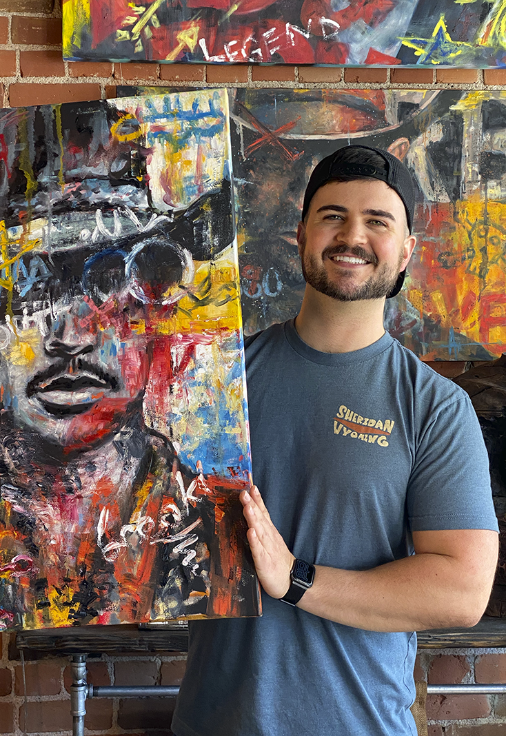 A smiling man in a blue t-shirt and black cap holding a colorful abstract portrait painting of a person with glasses and dark hair, set against a brick wall with other vibrant paintings in the background.