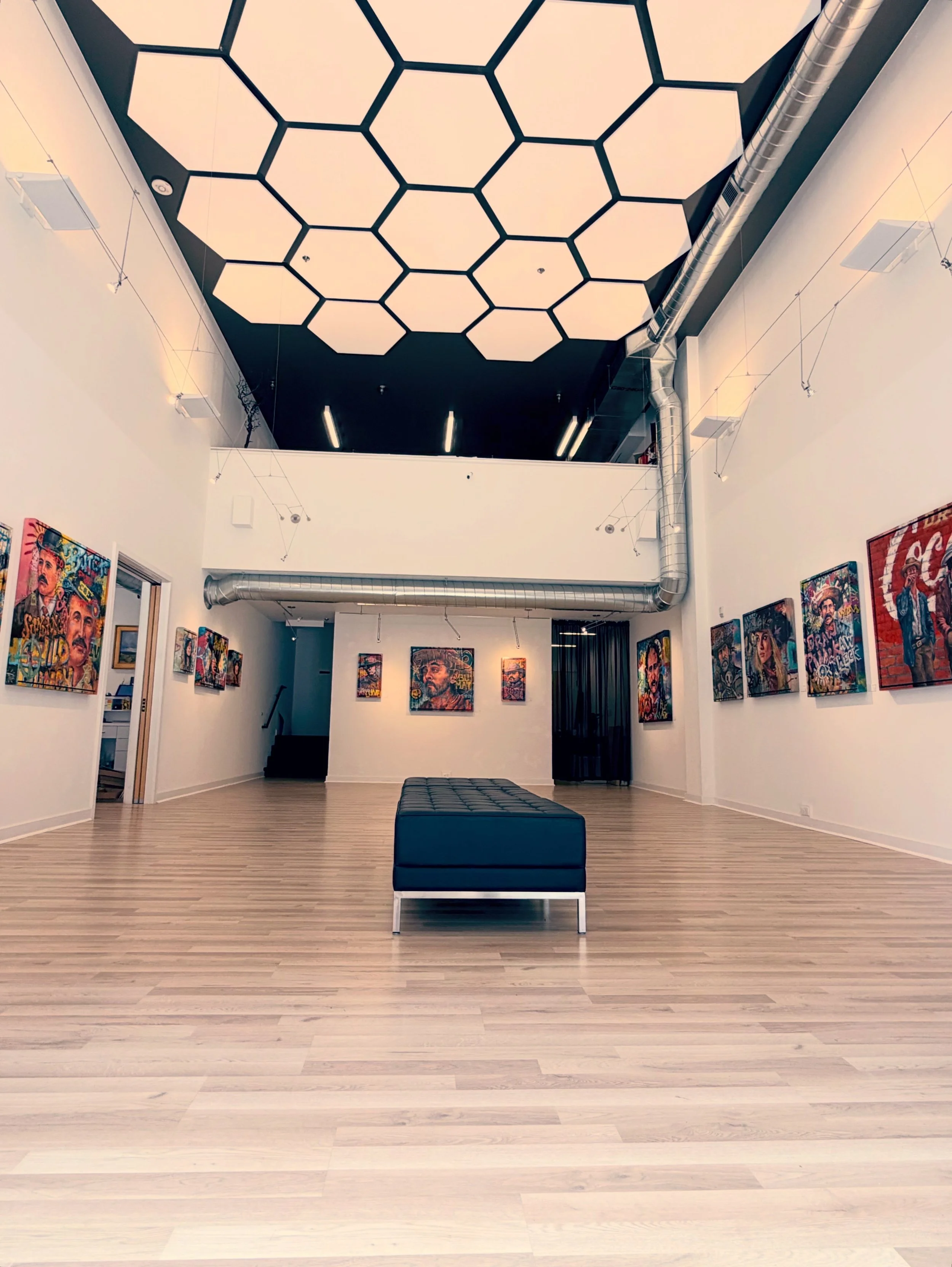 An art gallery with a high ceiling with large hexagon-shaped lights, artwork on the walls, a black cushioned bench in the center, and exposed silver ductwork.