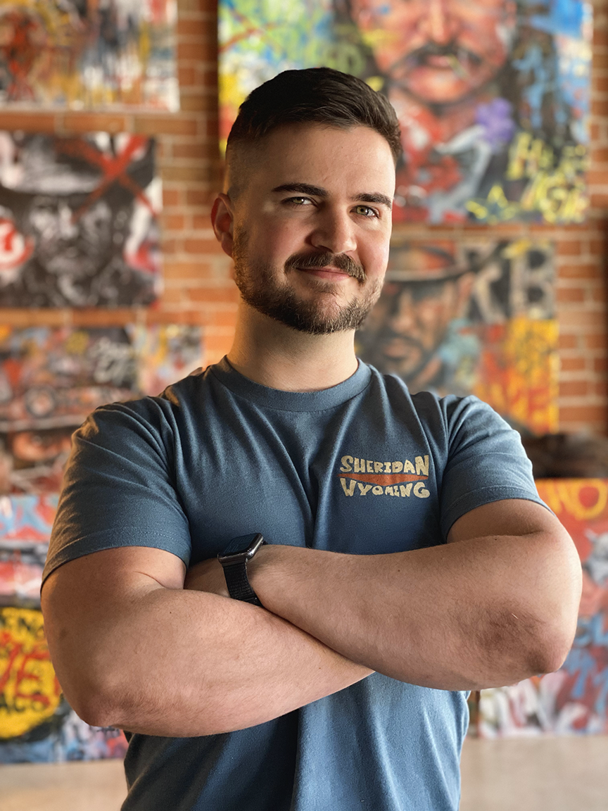 A young man with short dark hair and a beard, wearing a blue t-shirt with "Sheridan Wyoming" written on it, standing in front of colorful abstract artwork on a brick wall, with arms crossed and smiling.