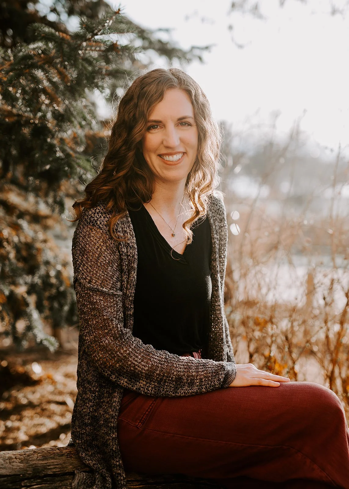 A woman with long wavy hair smiling outdoors near a tree with autumn foliage, wearing a black top, patterned cardigan, and maroon pants.