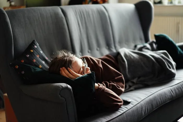 Person sleeping on a gray sofa with a black pillow and resting head on their hand