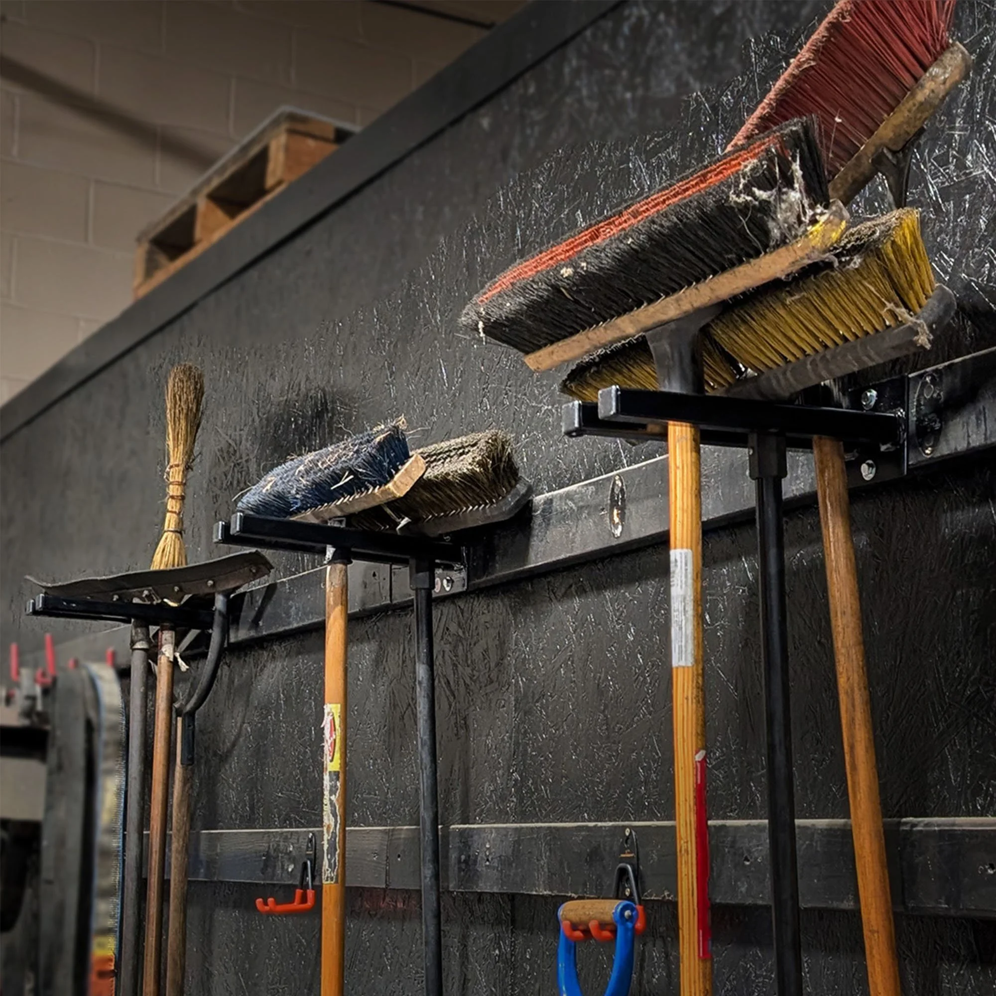 Double-prong tool racks mounted on shop wall holding brooms, rakes, and long-handled tools
