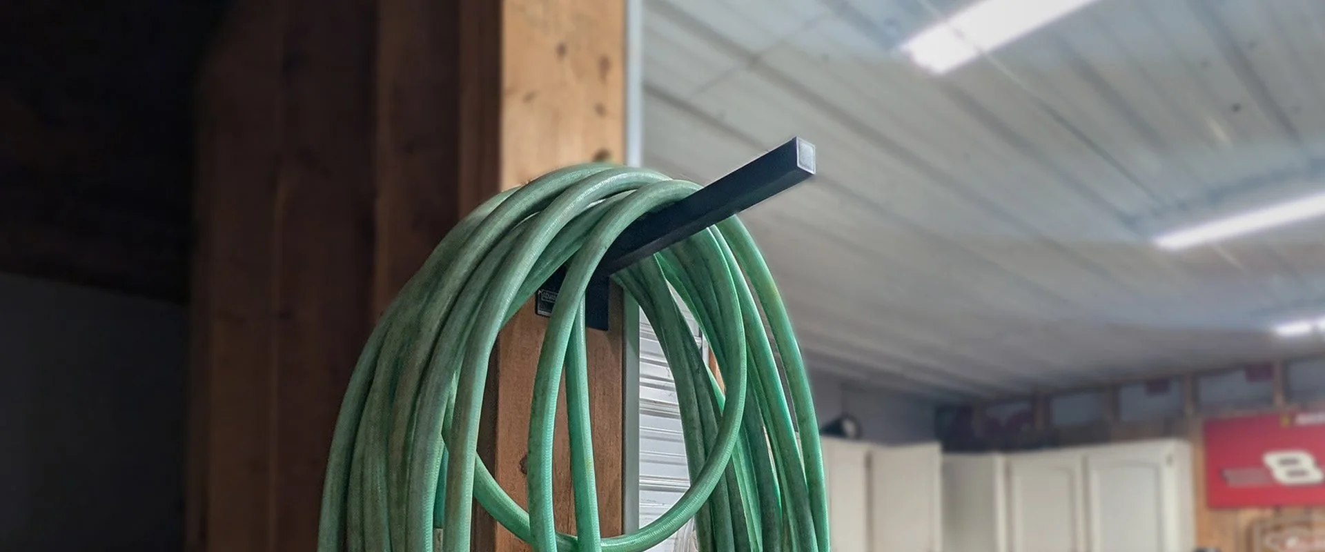 12-inch Single Prong Tool Rack in a Michigan workshop holding a coiled green garden hose, wall-mounted installation shown in a real working shop, powder-coated black 16-gauge American steel, made in Grand Rapids Michigan
